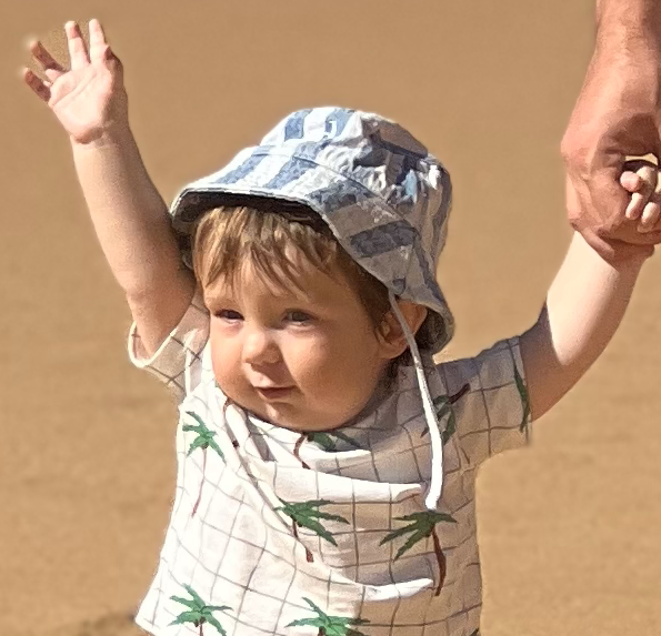 A young child wearing a blue and white sun hat and a shirt with palm trees, holding an adult's hand and walking outdoors on a sandy surface.