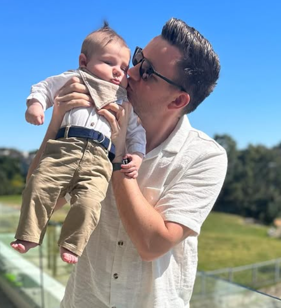A man holding a sleeping baby outdoors on a sunny day, with a grassy landscape and trees in the background.