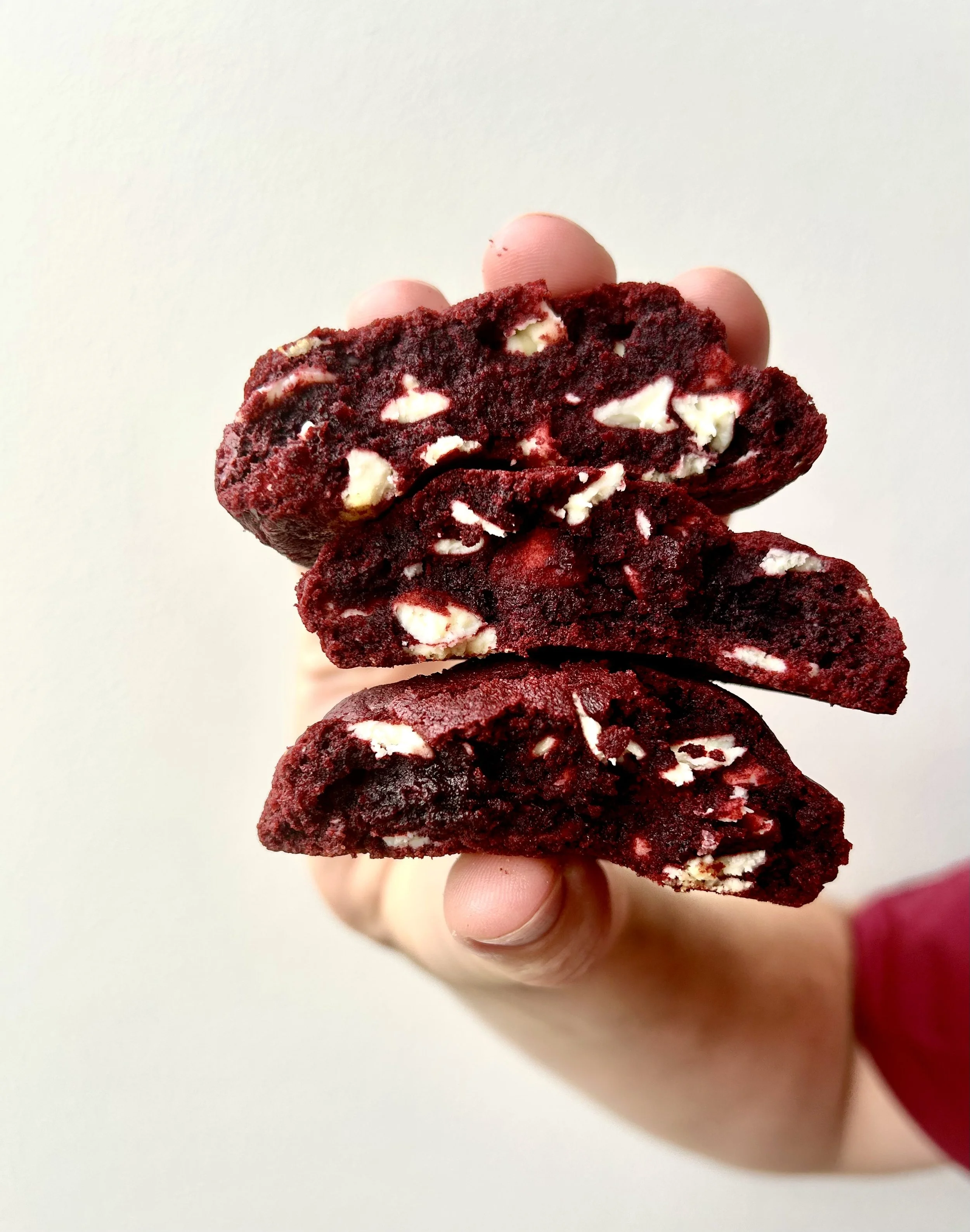 Hand holding three red velvet cookies with white chocolate chunks against a plain white background.