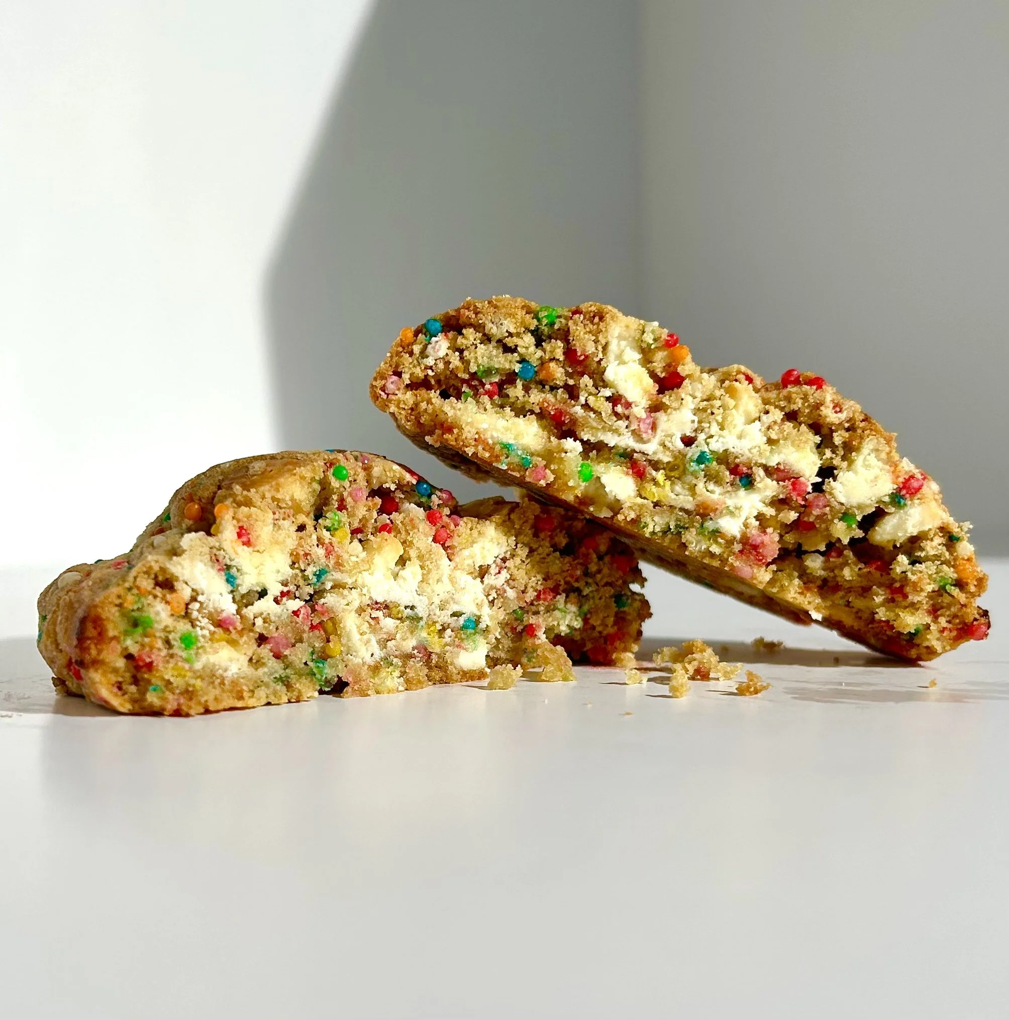 Two broken sugar cookies with colorful sprinkles on a white surface with a shadowed background.
