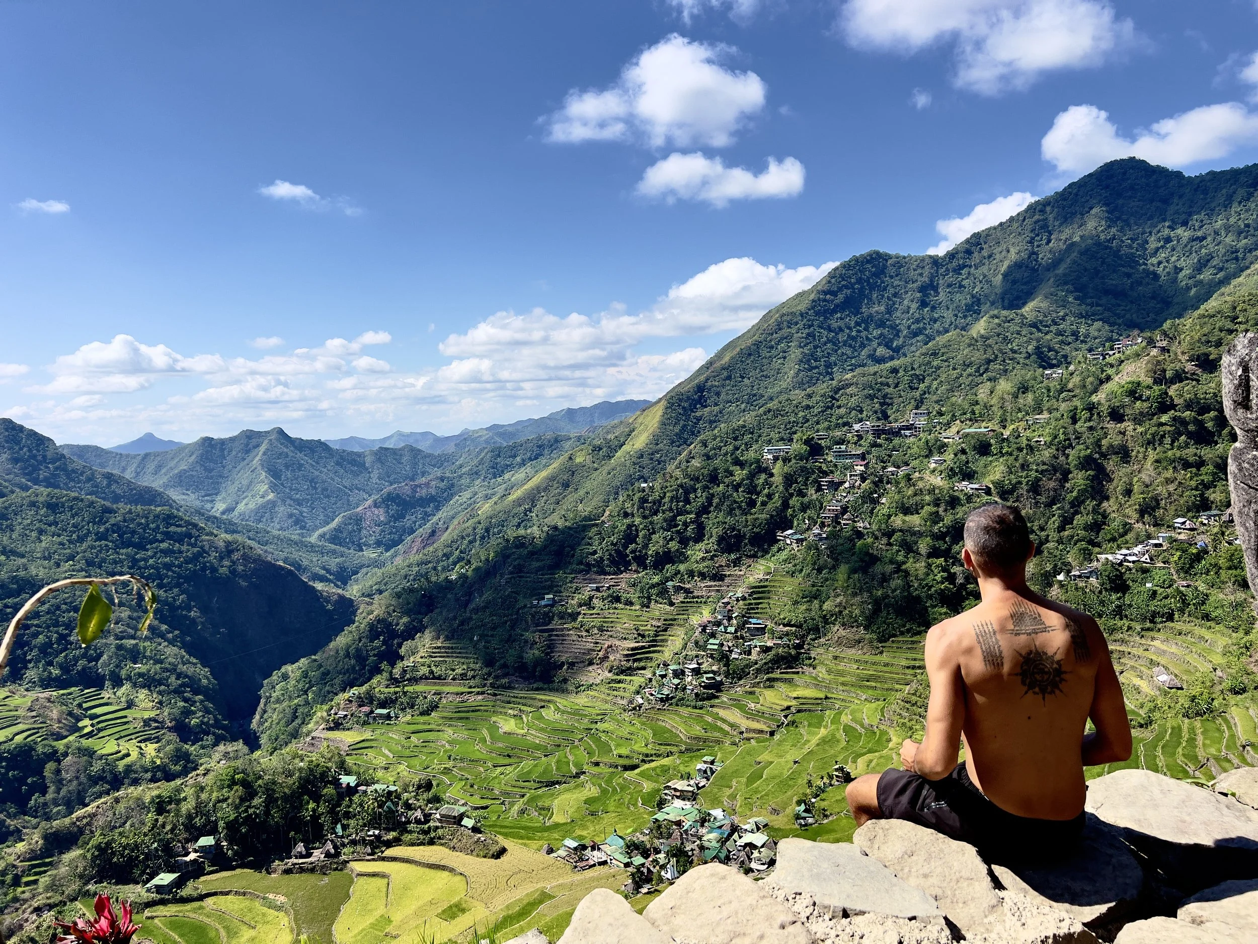 A shirtless man with tattoos sits cross-legged on a rocky ledge, overlooking terraced rice fields and green mountains under a partly cloudy blue sky.
