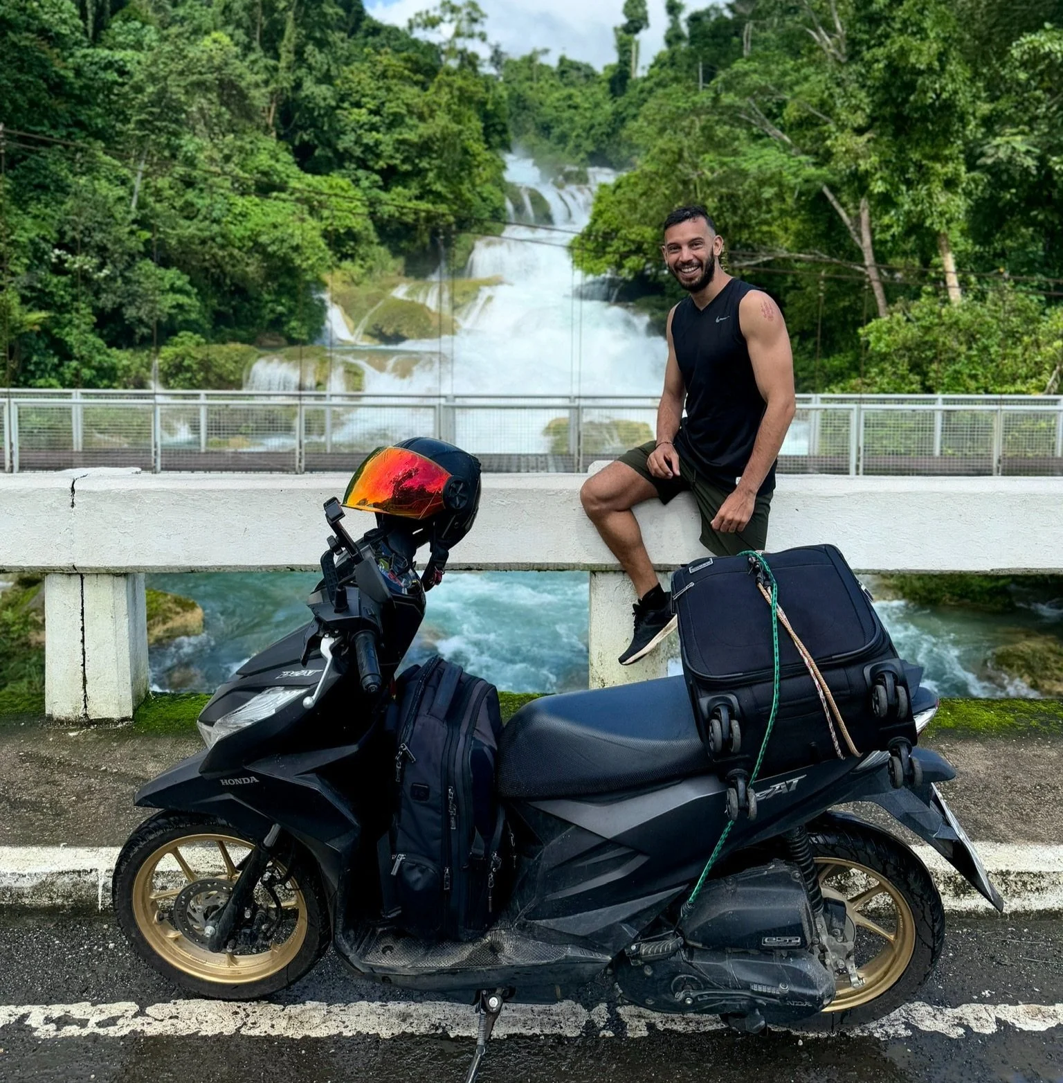 A smiling man sitting on a bridge ledge next to a black motorcycle with a red helmet on the handlebars, a backpack and a bag attached, overlooking a waterfall and lush green trees.