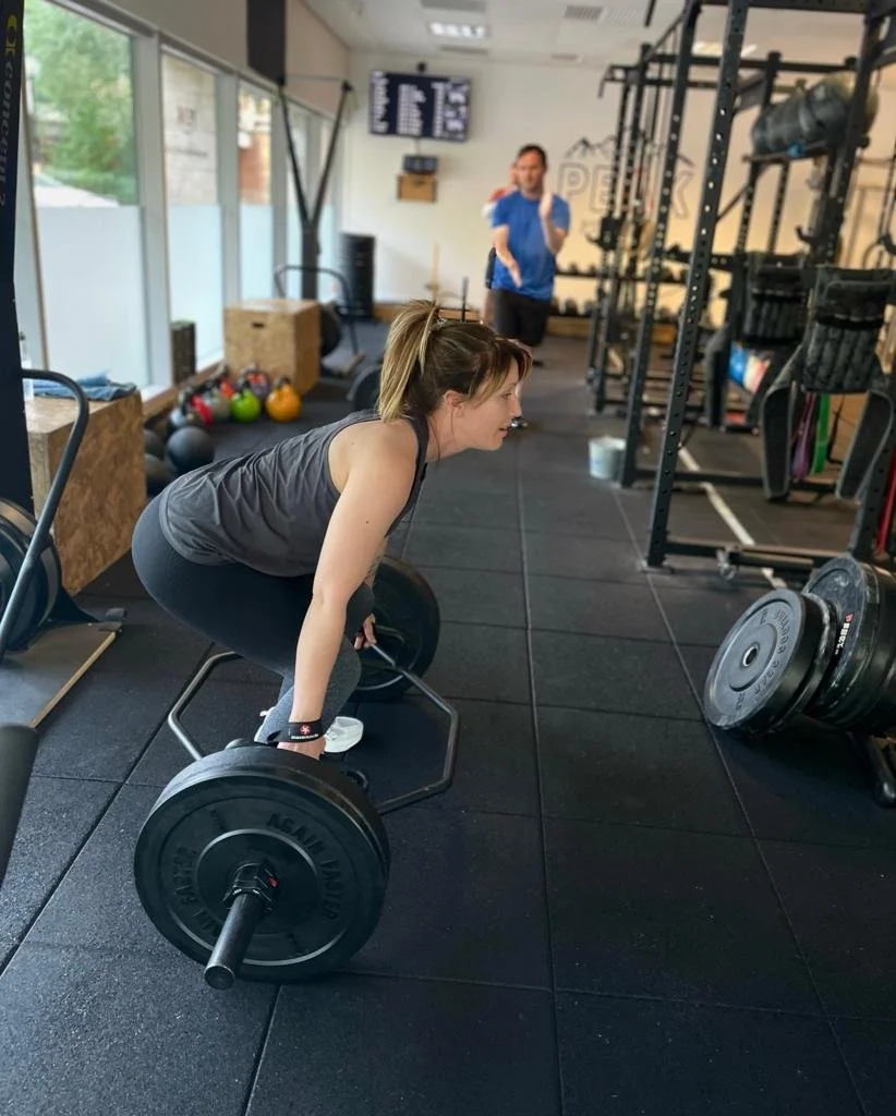 A woman performing a deadlift exercise in a gym while a man in a blue shirt stands in the background.