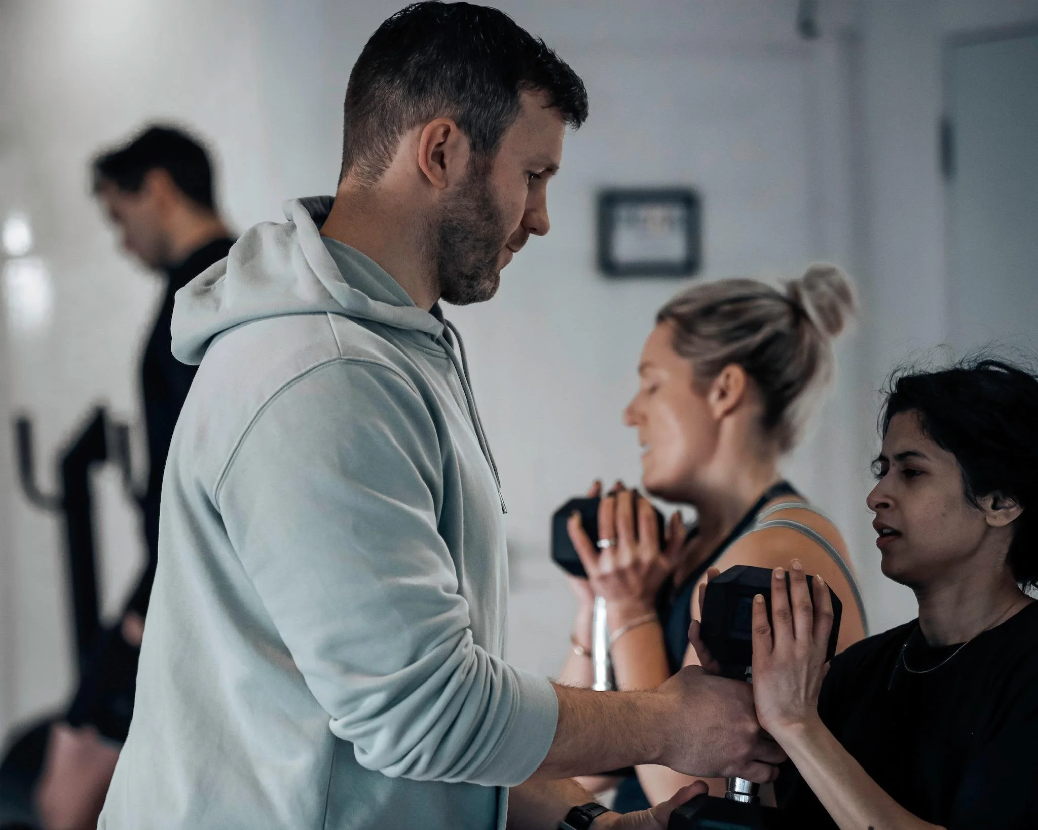A man in a gray hoodie leaning over and holding hands of a woman with dark hair and a black stop sign tattoo, who is wearing a dark shirt. The scene appears to be a medical or clinical setting with other people in the background.
