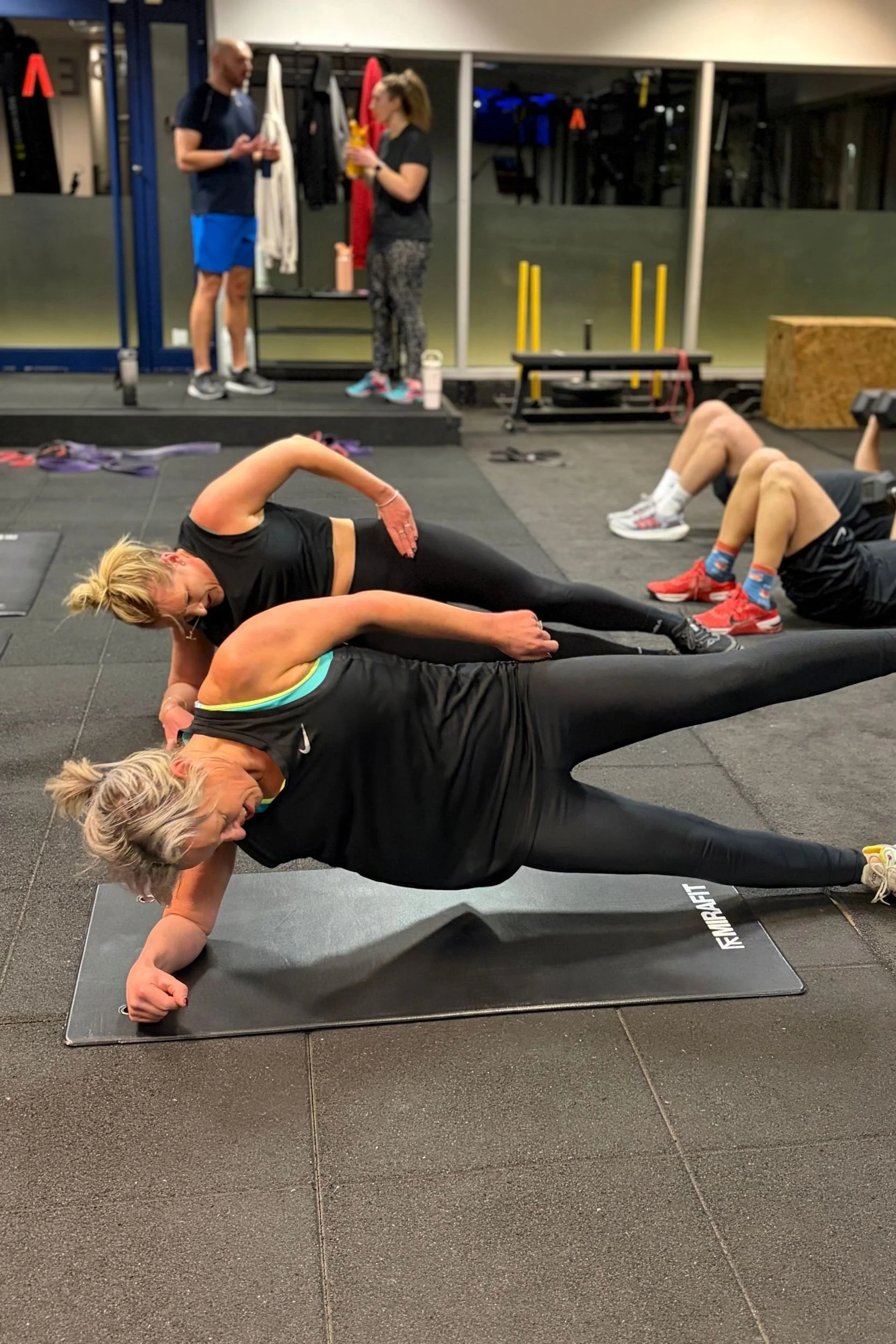 Women doing floor exercises on mats in a gym, with two people standing and talking in the background.