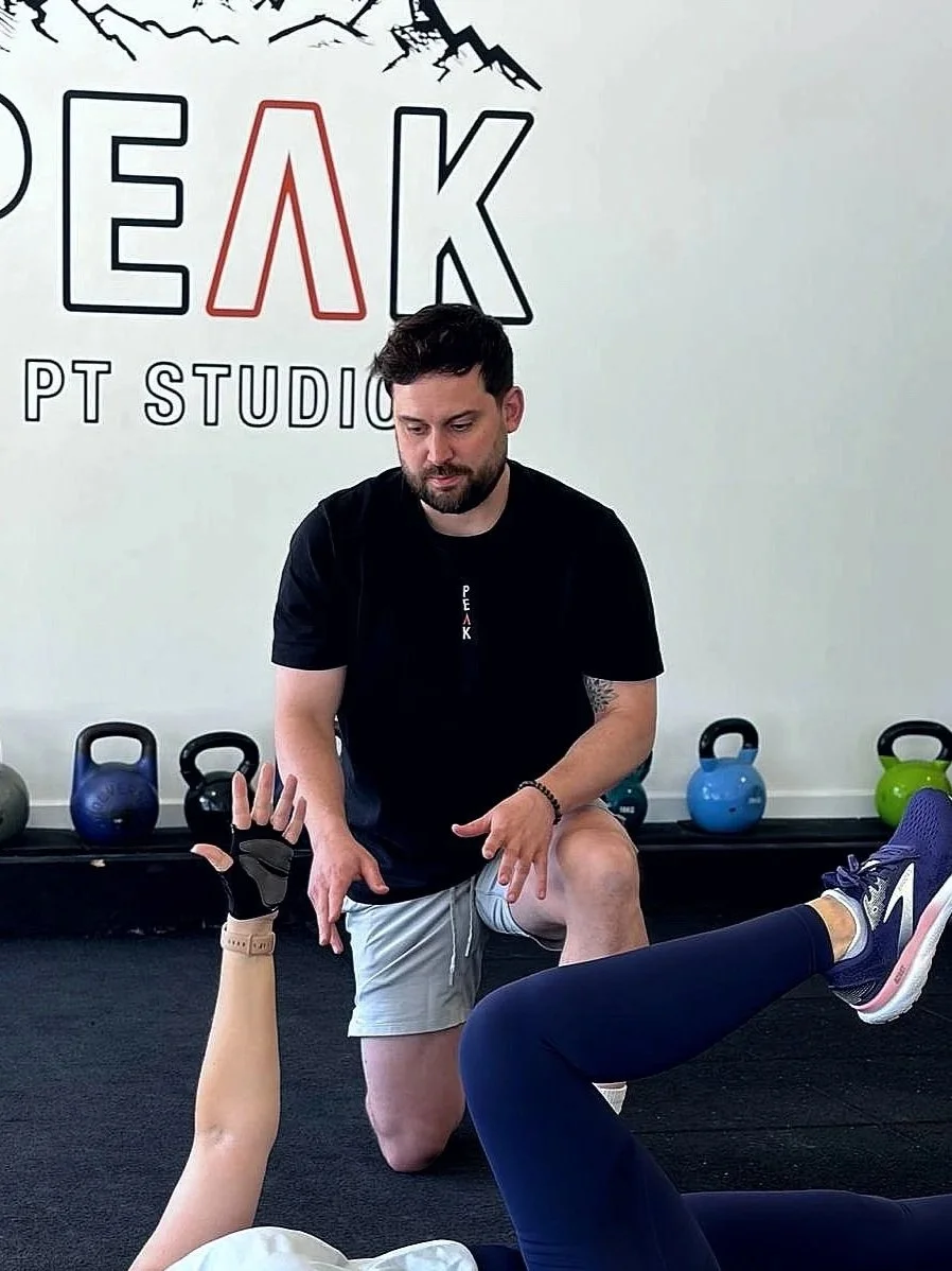 A trainer kneeling in front of a person lying on the floor, assisting with a workout at Peak PT Studio, with kettlebells on the wall behind them.