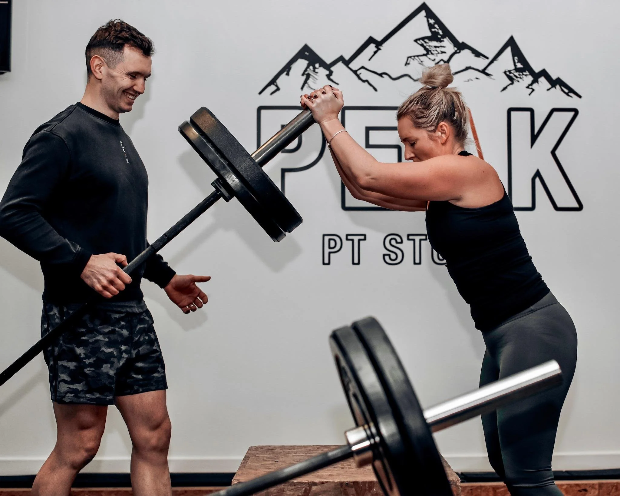 A woman is arm wrestling a man at a fitness gym while a large weightlifting barbell and weights are in the foreground. They are smiling and appear to be enjoying the competition.