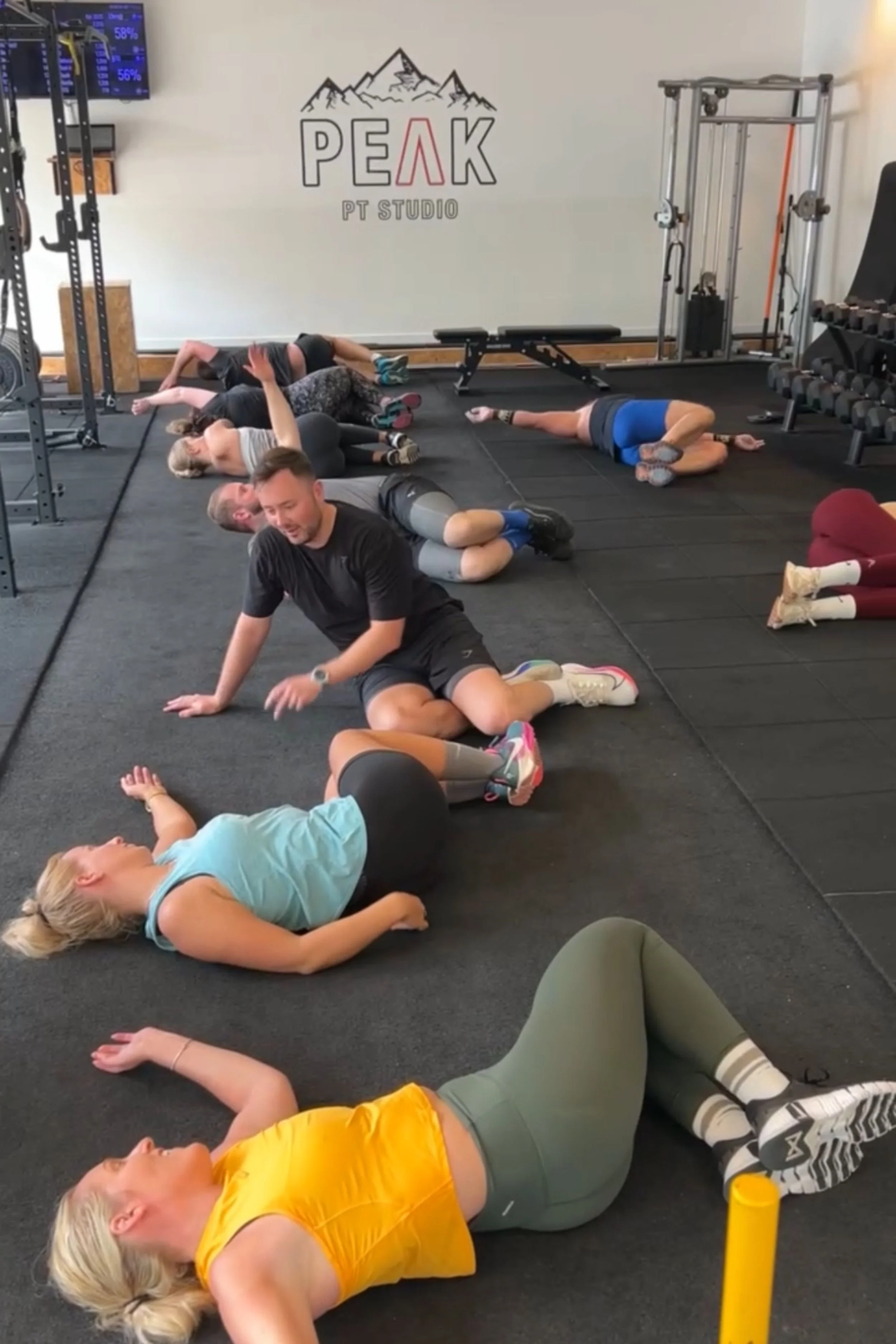 Group of people performing floor exercises or stretching in a gym with a logo on the wall reading 'PEAK PT Studio' and mountain graphic, fitness equipment around.