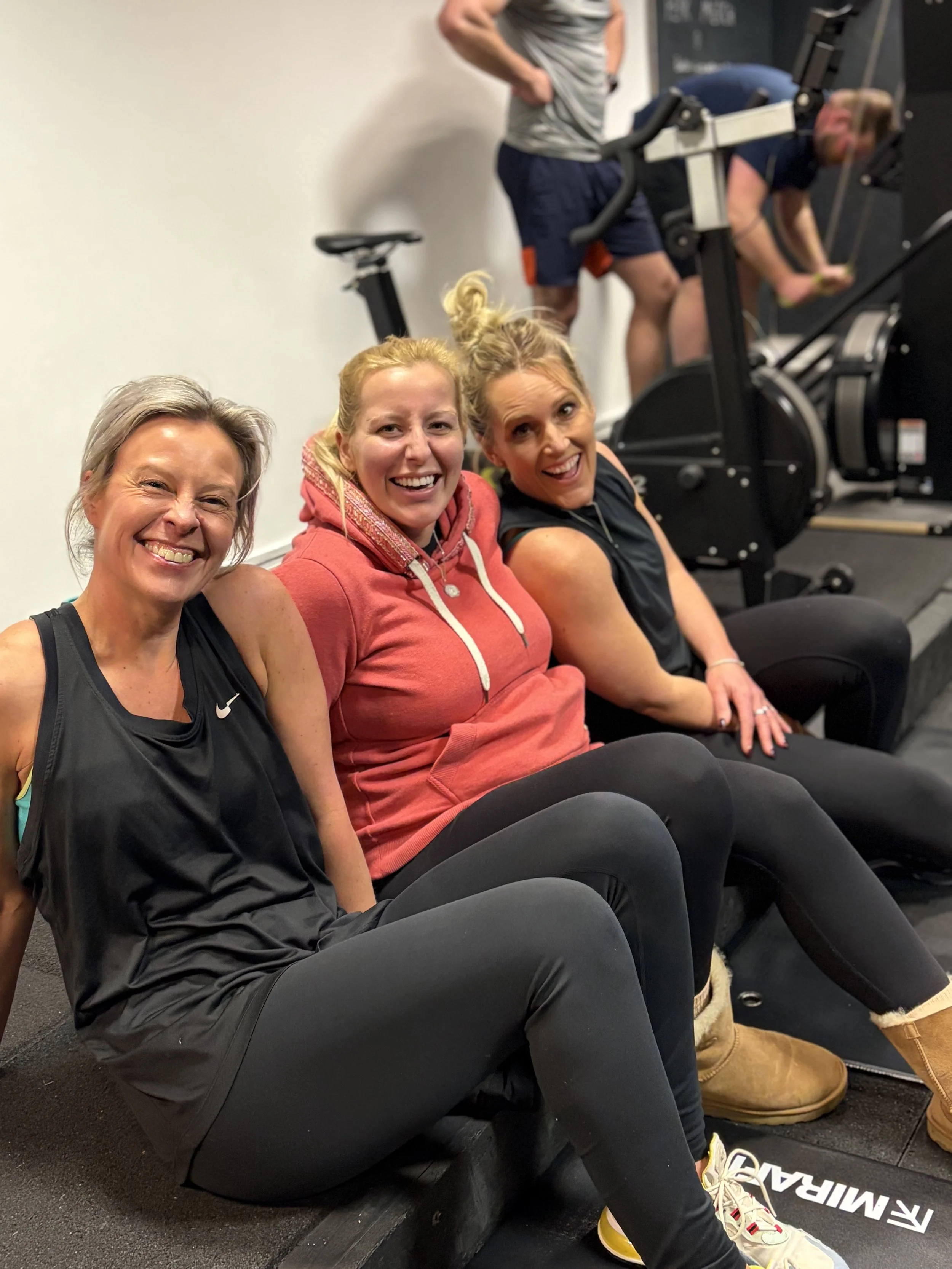 Three women sit on the floor at a gym, smiling at the camera, with fitness equipment and two men working out in the background.