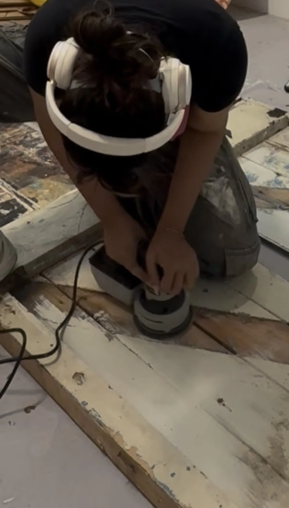 Person kneeling on the floor working on a wooden surface with a power tool, wearing a black shirt, camouflage pants, and white headphones.
