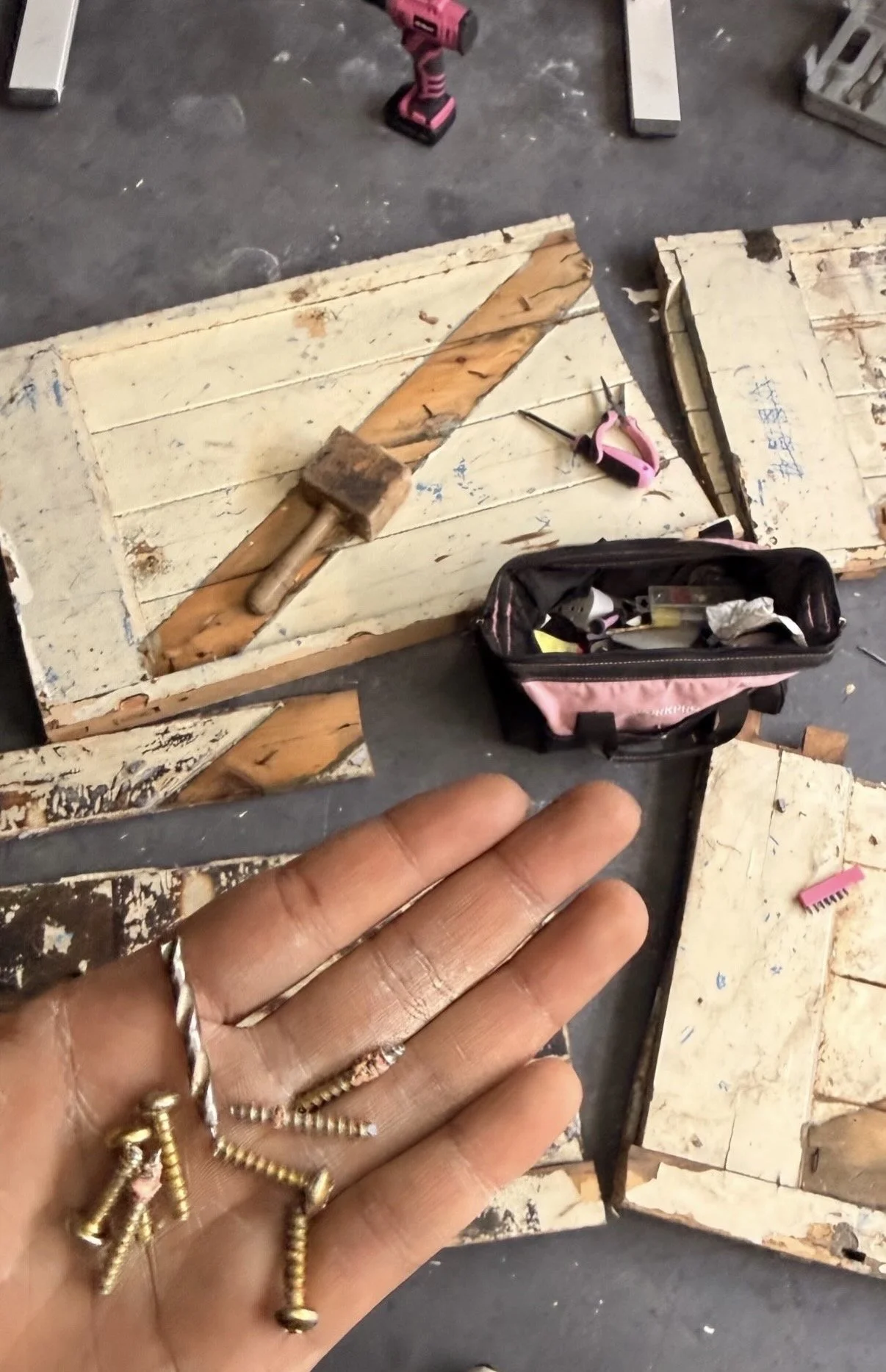 A person's hand, showing nails, resting in front of a woodworking workspace. On the work surface, there are pieces of aged, painted wood, a pink cordless drill, a pink hand tool, and a black and pink toolbox with various tools inside.
