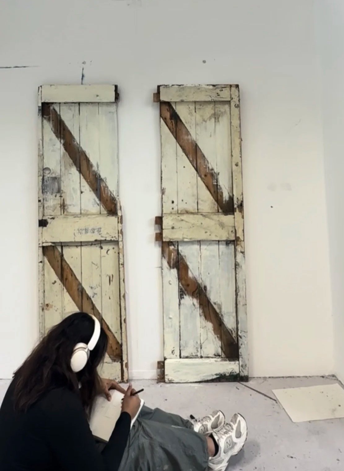 A girl with headphones sitting on the floor, writing in a notebook while leaning against a wall with two rustic, weathered wooden doors.