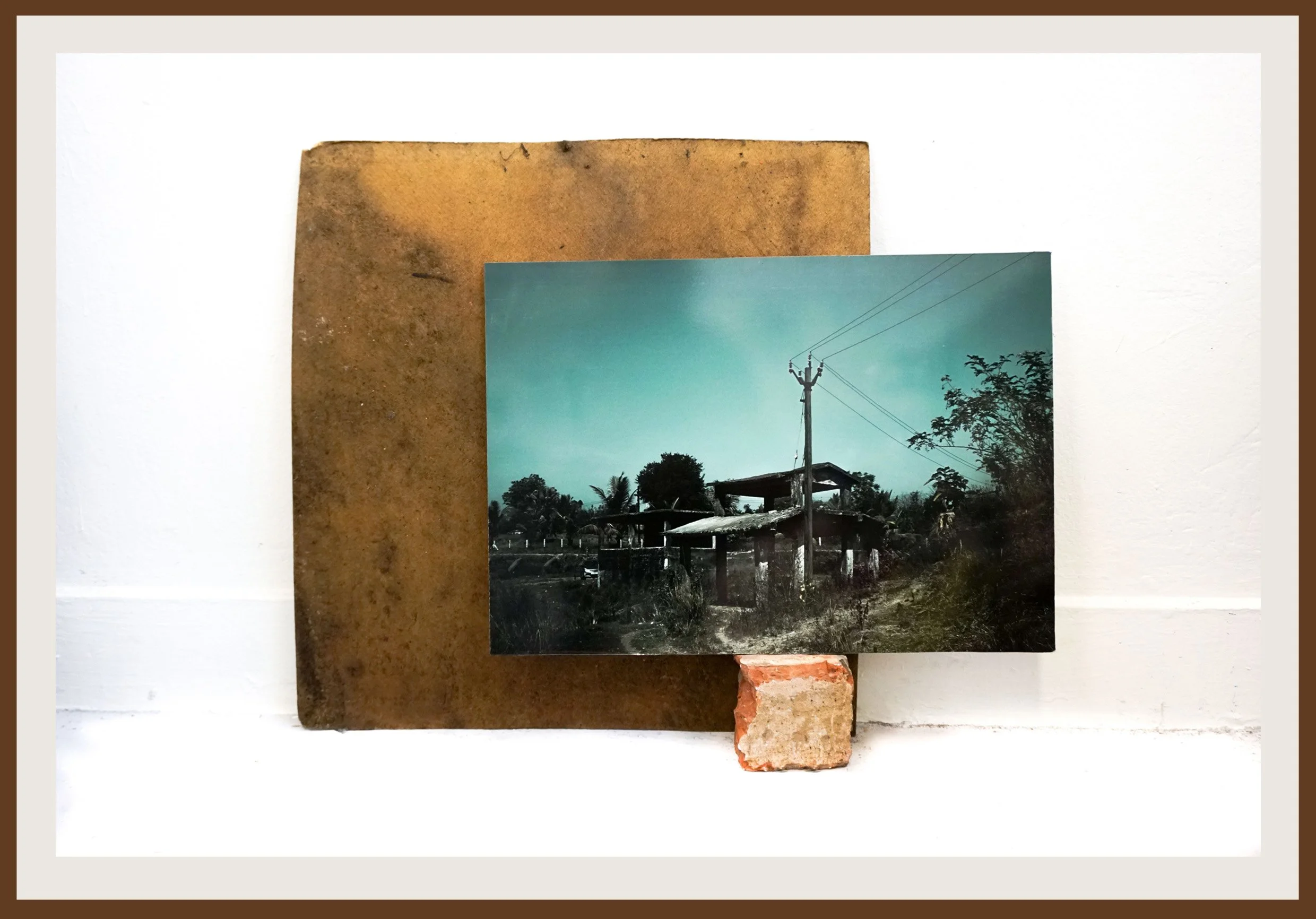 A photograph of a rural area, power lines, and houses, mounted on a brown board, with a small brick at the bottom right corner.