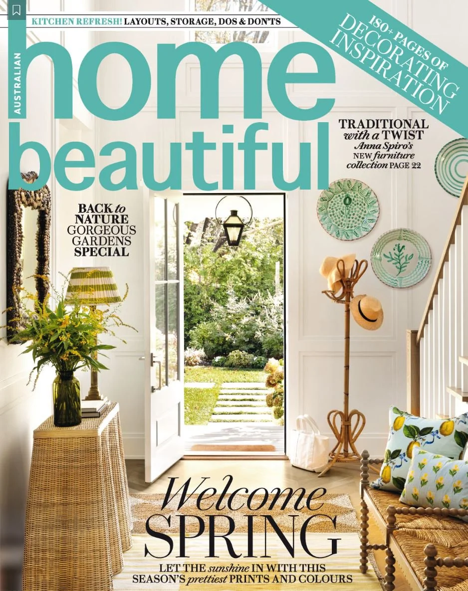 Cover of 'Home Beautiful' magazine featuring a bright, inviting entrance hall with a door open to a garden, decorated with wall art, cushions, a wicker table with a vase of flowers, and a coat rack with hats.