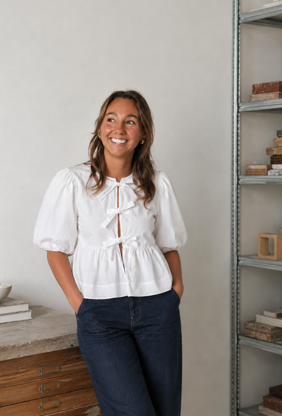 A woman with brown hair and earrings smiling, wearing a white puff-sleeve blouse with bow details and dark jeans, standing indoors near a metal shelf and a wooden dresser.