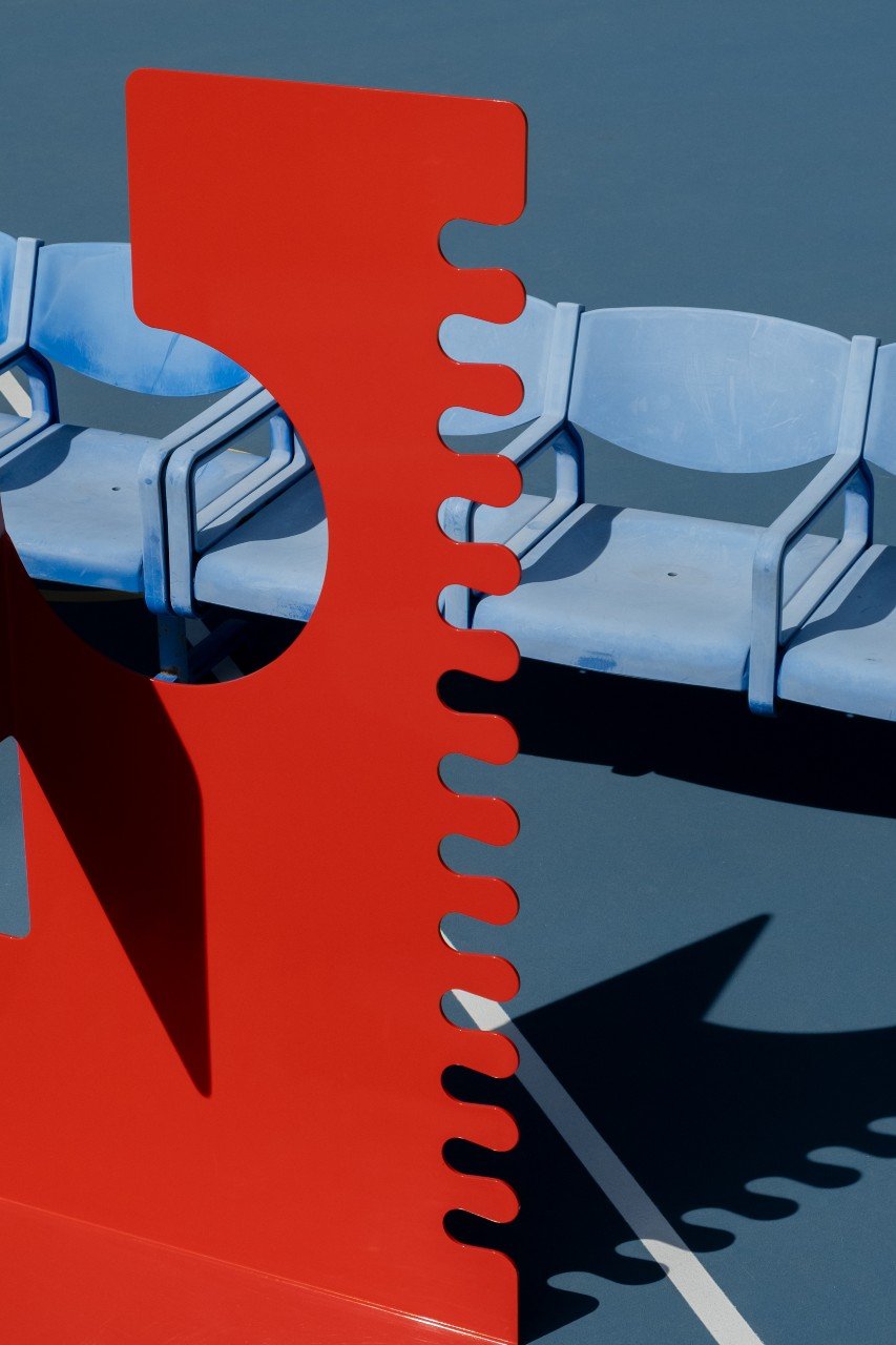 Standing Signal Red Sculpture, powder coated aluminium sheet, photographed in front of blue bleacher seats, on a blue tennis court