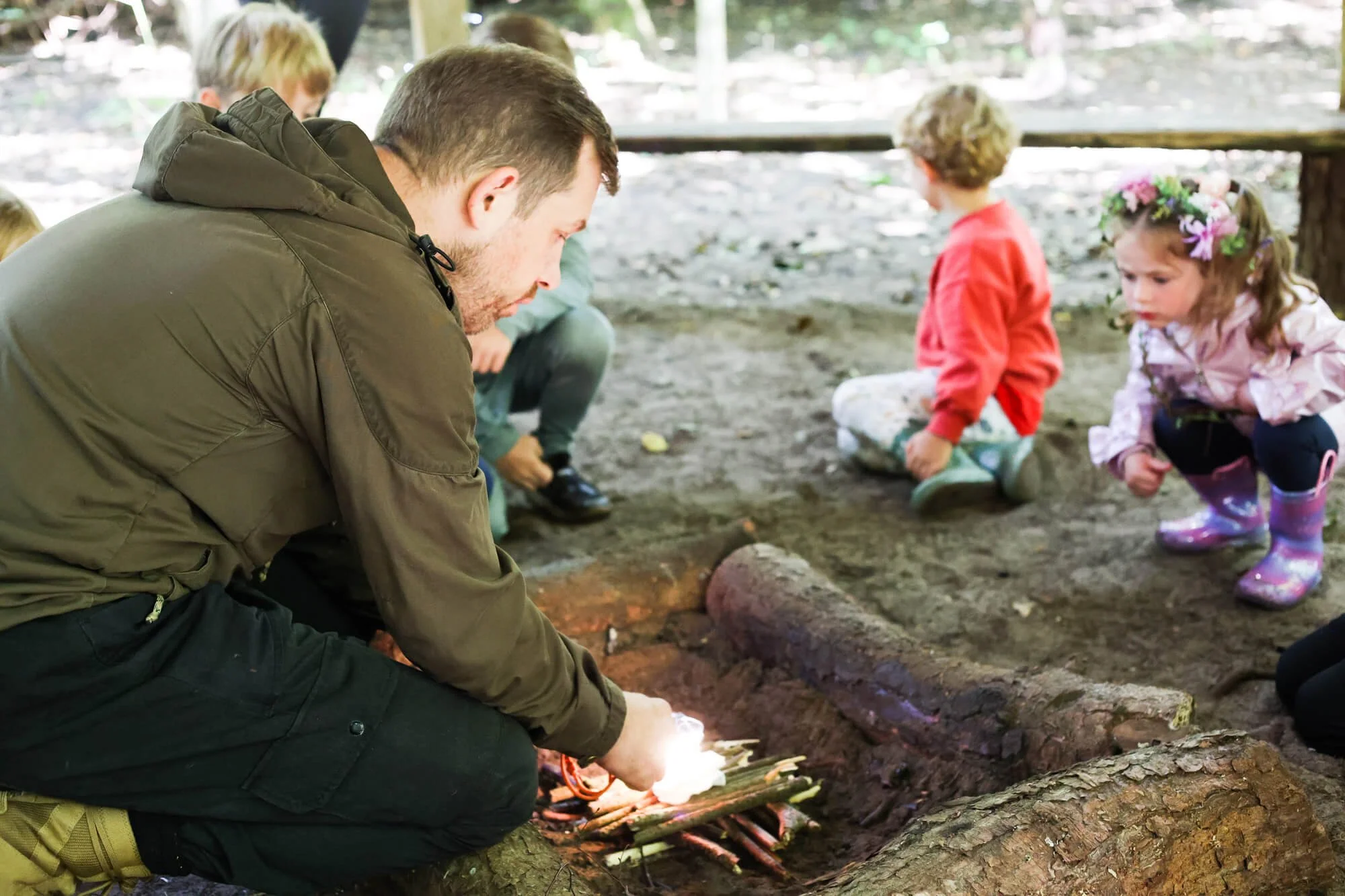 A man in a brown jacket crouches and lights a campfire with a small flame in a forested outdoor area, while children sit nearby observing.