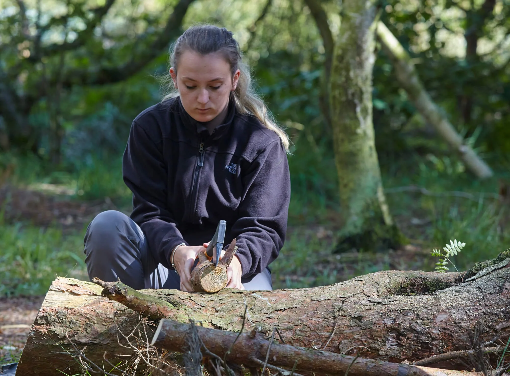 A woman with long hair tied back, wearing a black jacket, is chopping a log with an axe in a forest.
