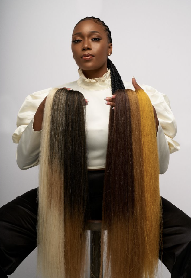 A woman holding two long hair extensions, one blonde and one brown, while sitting against a plain background.