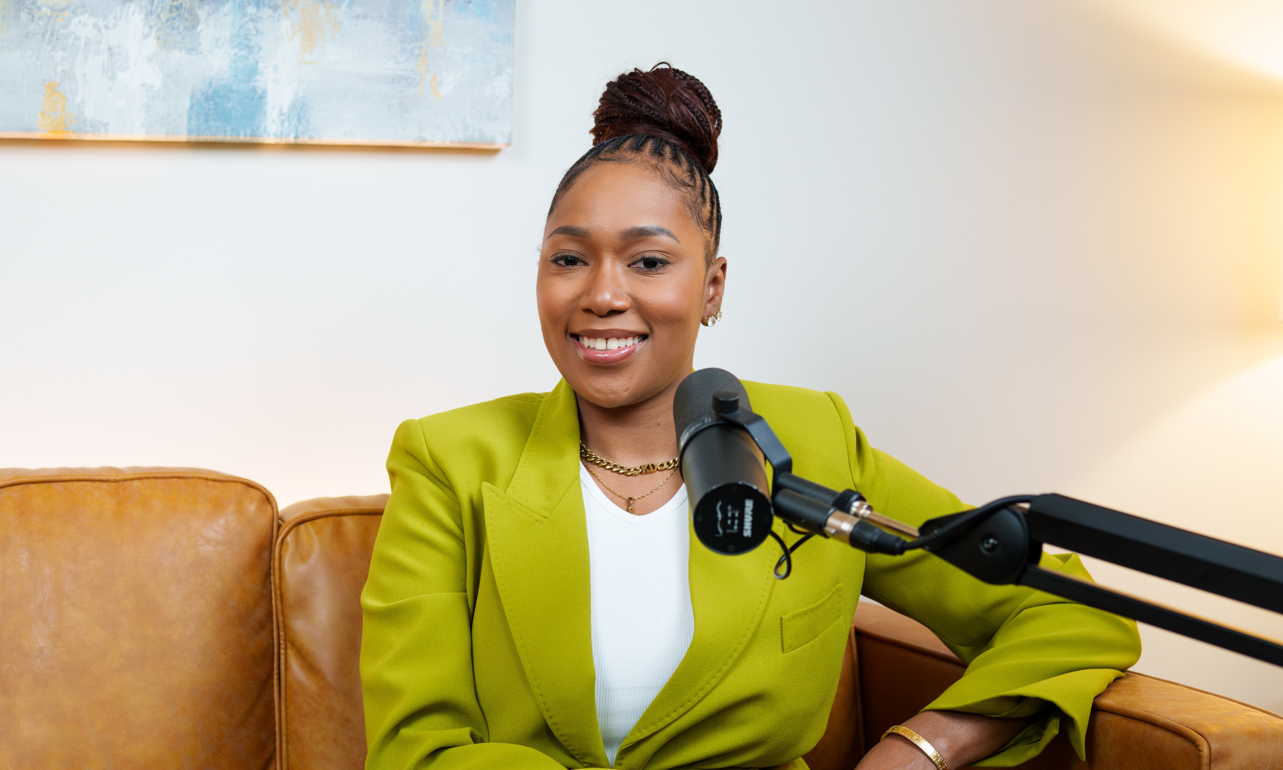Woman with red dreadlocks recording a podcast or video call using a microphone, sitting on a pink couch, with a laptop on a round table in front of her.