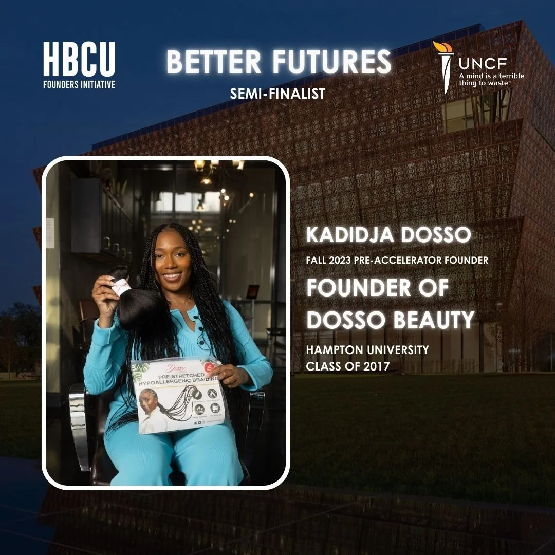 A woman smiling and holding a box and a hair product, sitting in a chair indoors. The woman is wearing a blue outfit and has long braided hair. The background shows a modern industrial-style space with lights and windows.