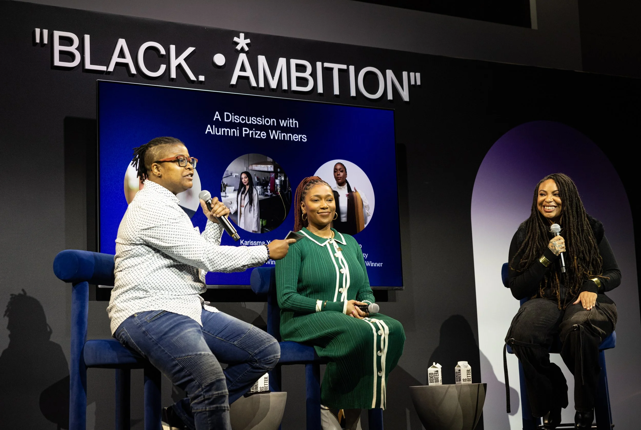 Panel discussion with three women on stage, two holding microphones, seated in front of a large screen displaying images of alumni and the text "A Discussion with Alumni Prize Winners" at an event titled "Black Ambition."