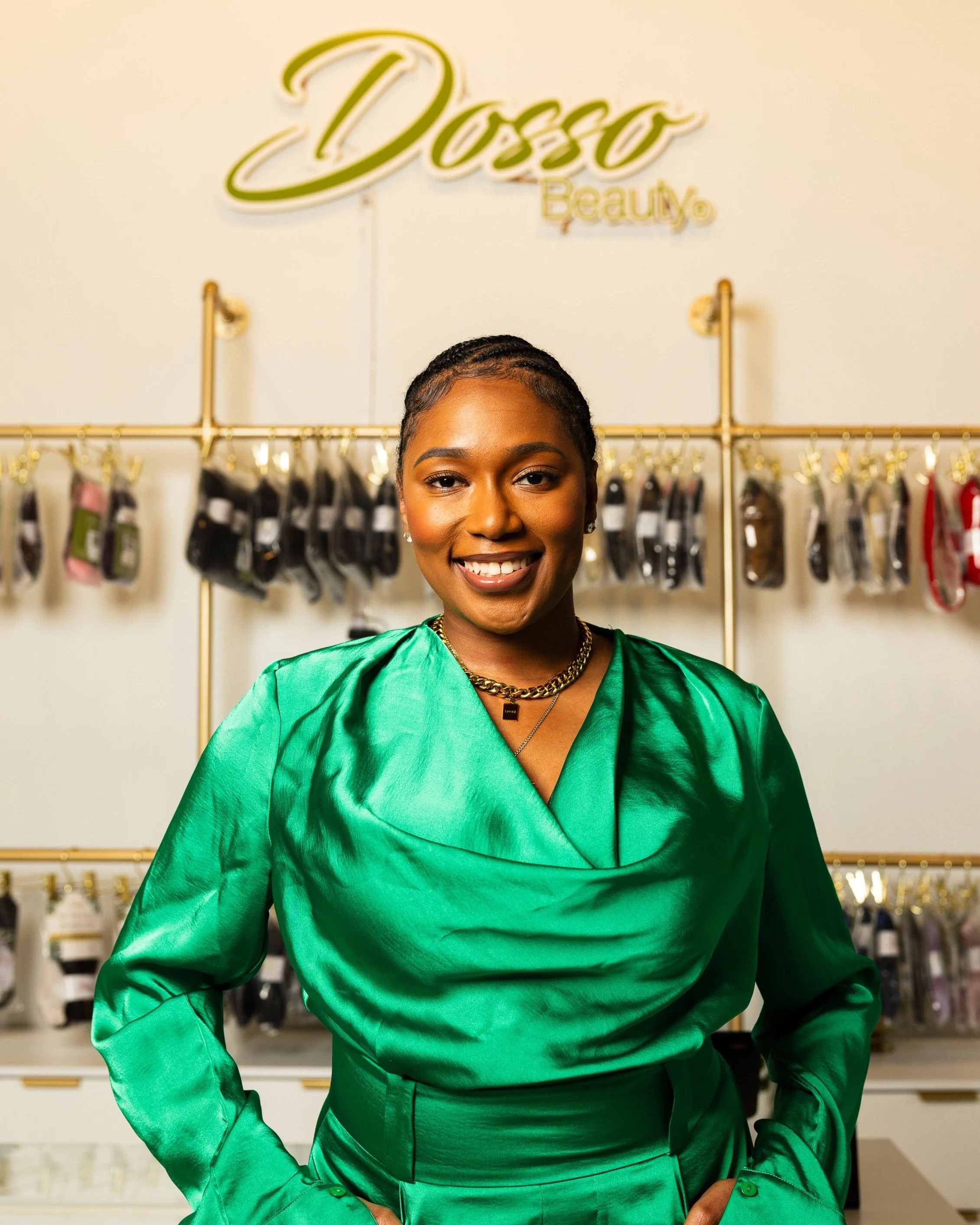 Smiling woman in a green satin dress standing inside a beauty store with jewelry and beauty products on display, behind a sign that says "Dosso Beauty."