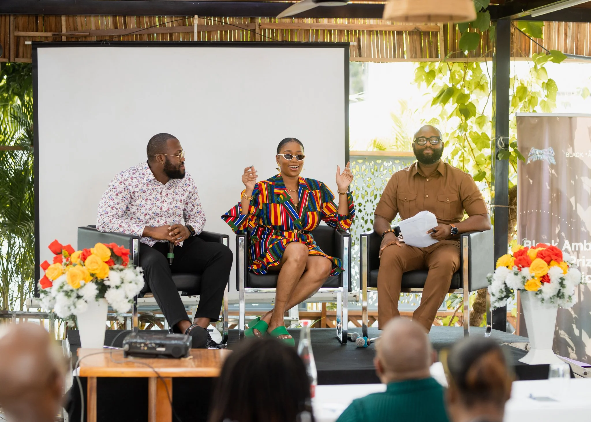 Three people speaking on a panel at an outdoor event, with a blank white screen behind them and audience members in front.