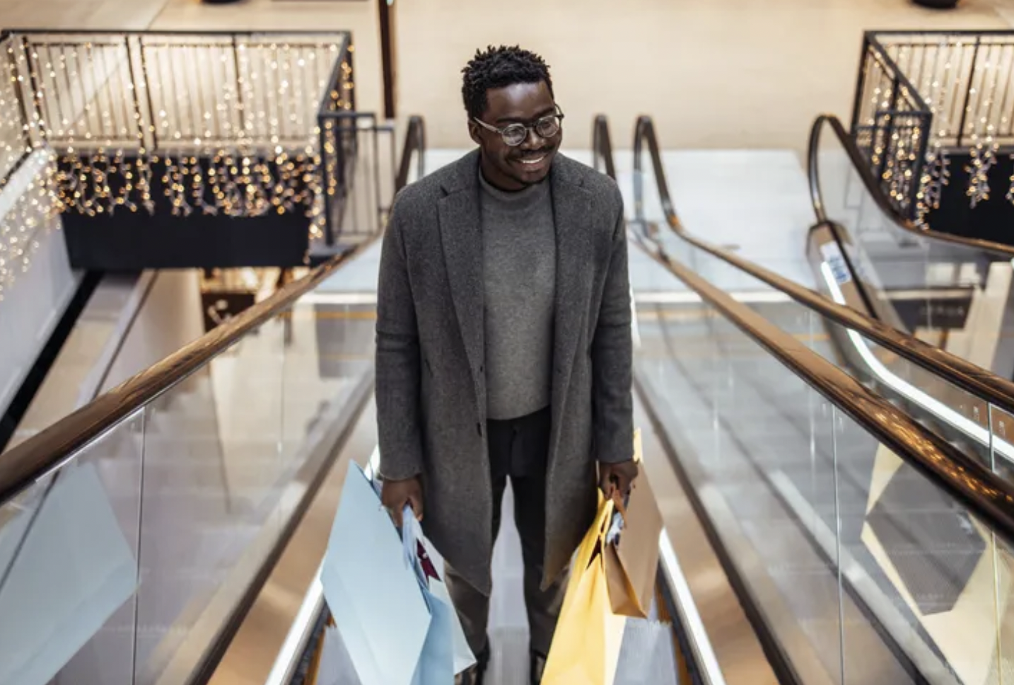 A man in a gray coat and glasses smiling while carrying shopping bags on an escalator in a mall decorated with string lights.
