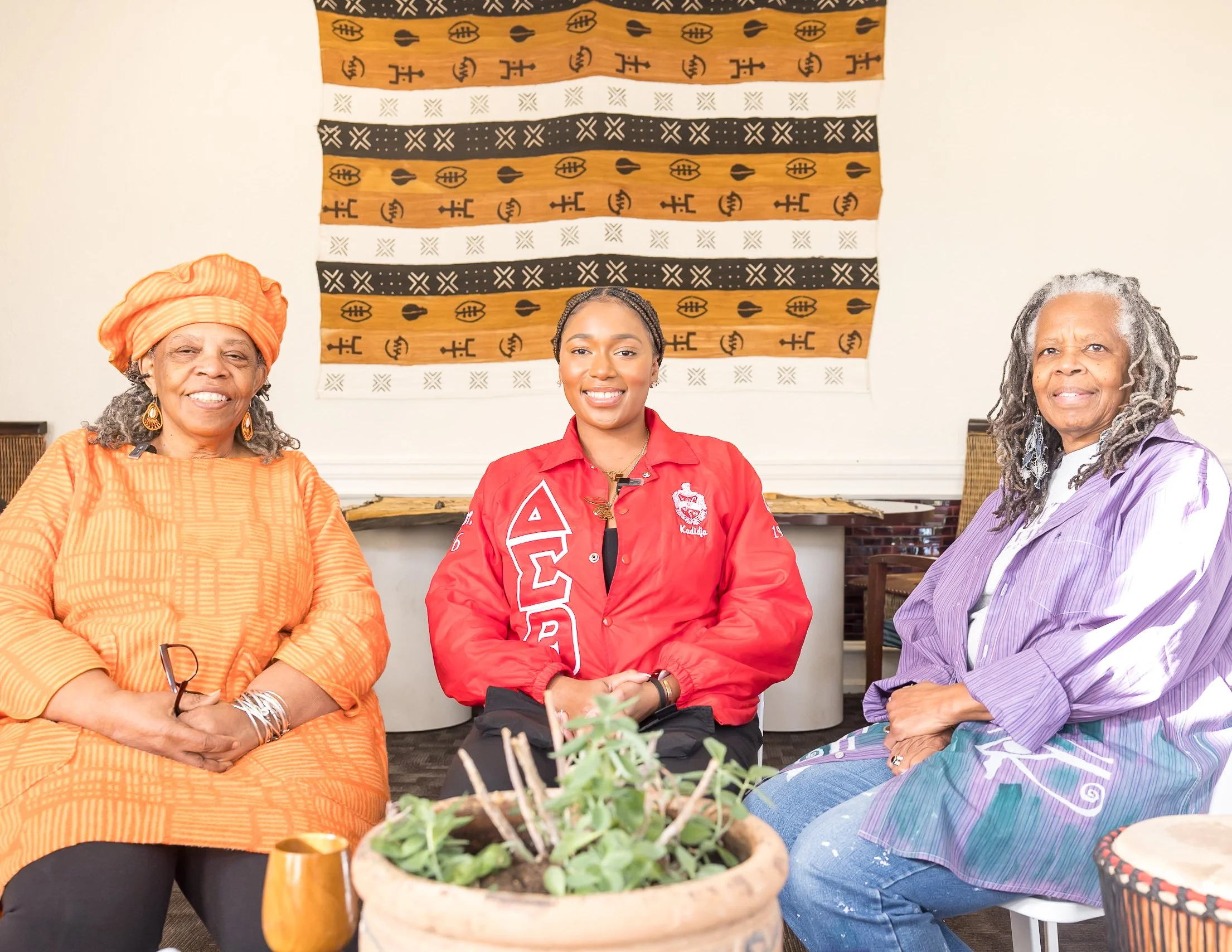 Three women sitting together indoors, smiling at the camera, with a decorative textile hanging on the wall behind them and a potted plant in the foreground.