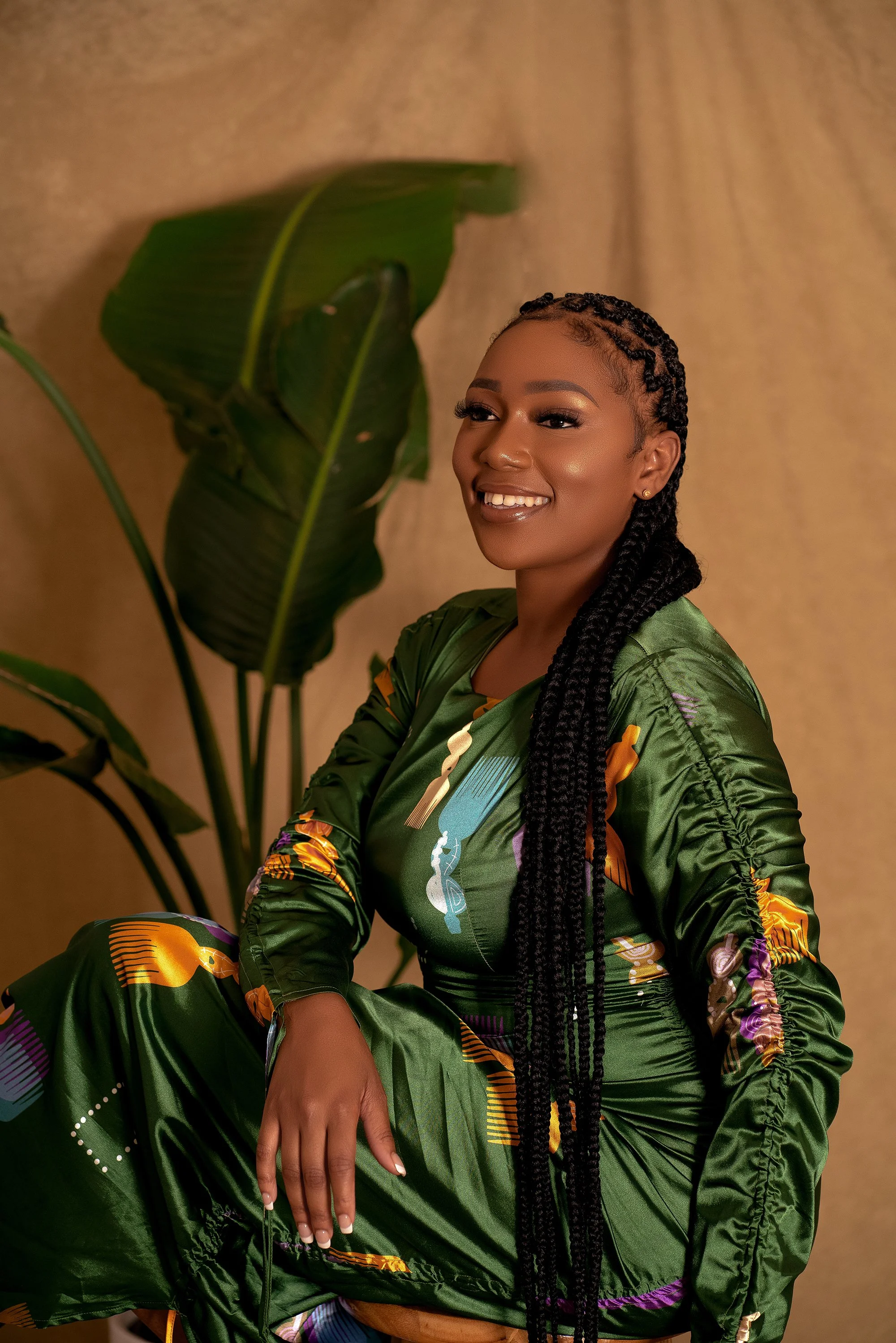 A woman with long braided hair smiling, dressed in a green patterned dress, sitting in front of a background with a large green tropical leaf.
