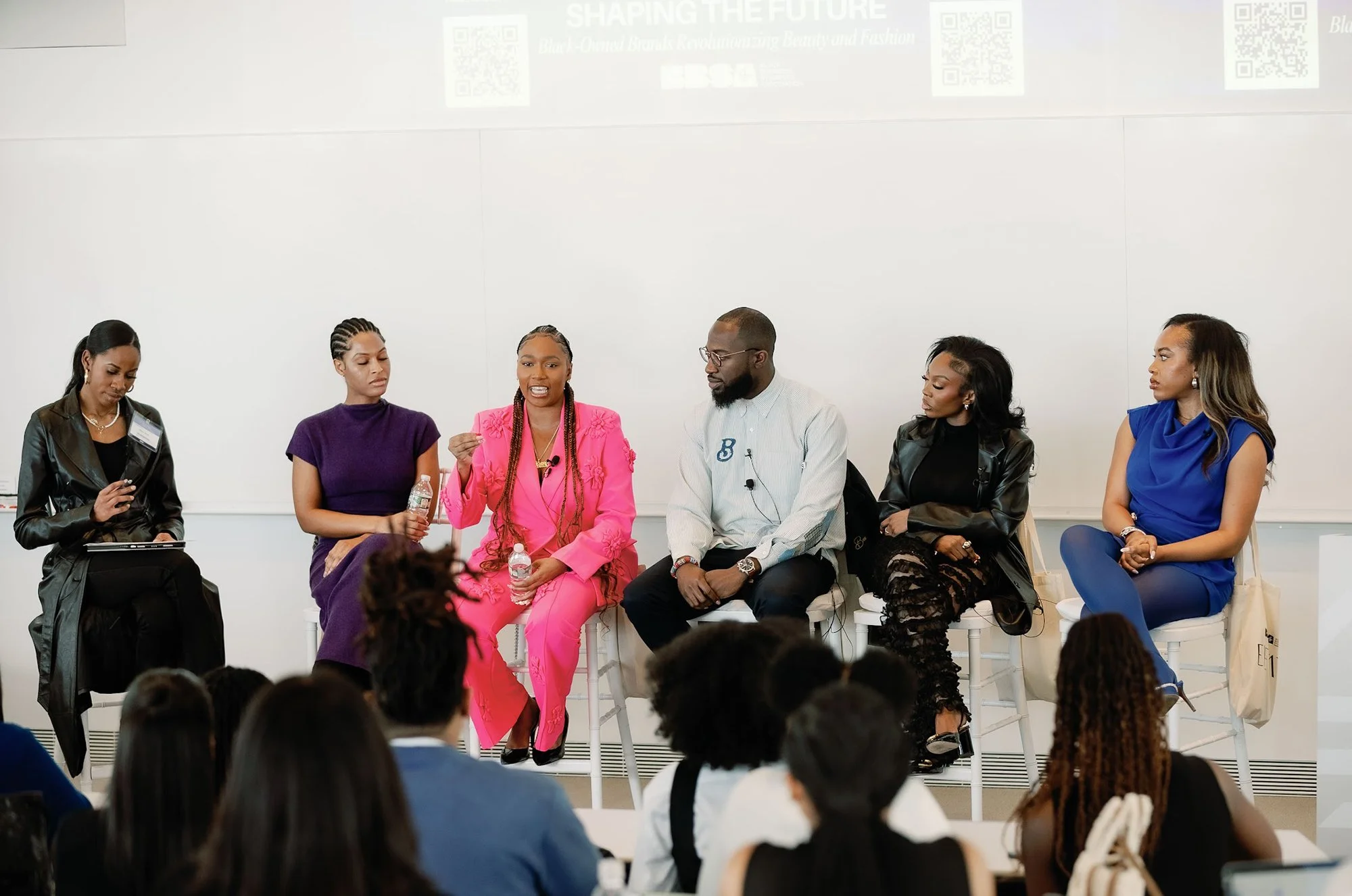Panel of six diverse individuals seated on a stage during a discussion, with an audience watching in front. The panelists include five women and one man, dressed in professional attire, with two women holding bottled water. A whiteboard or wall is behind them, displaying some text and QR codes.