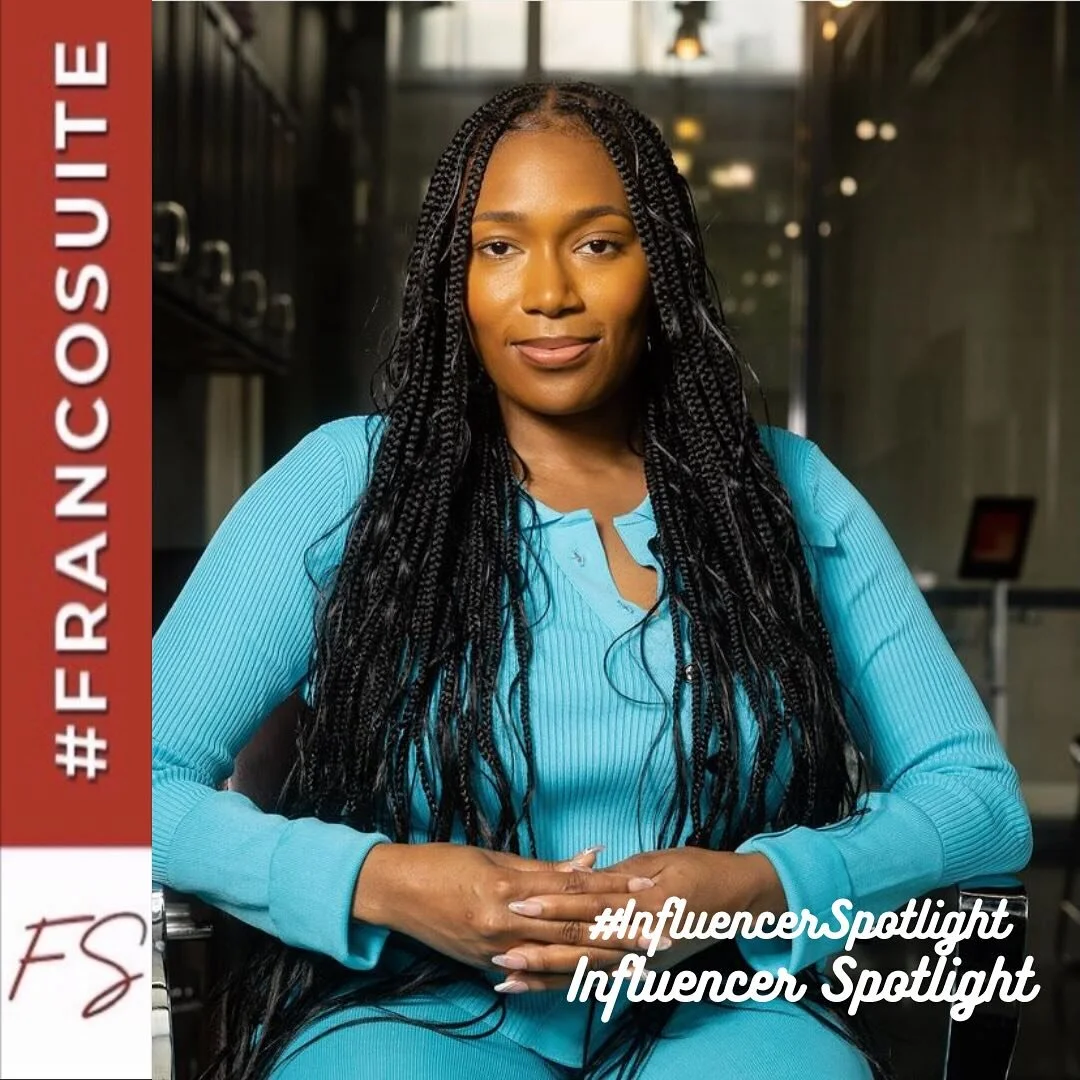 A woman with long braided hair sitting with her hands clasped in front of her, wearing a blue long sleeve shirt, in a modern indoor setting.