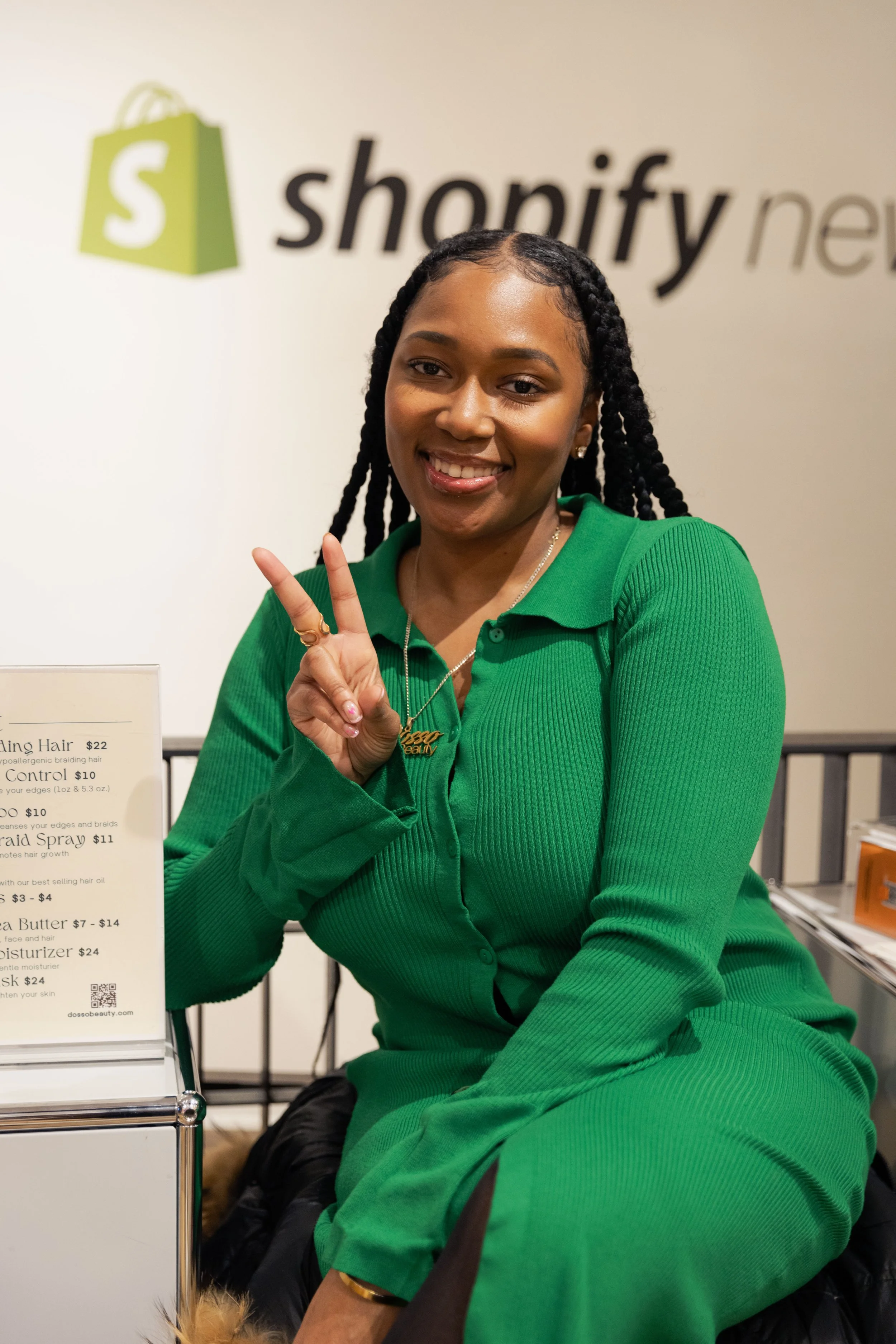 A woman with braided hair smiling and making a peace sign, sitting in front of a Shopify logo background.