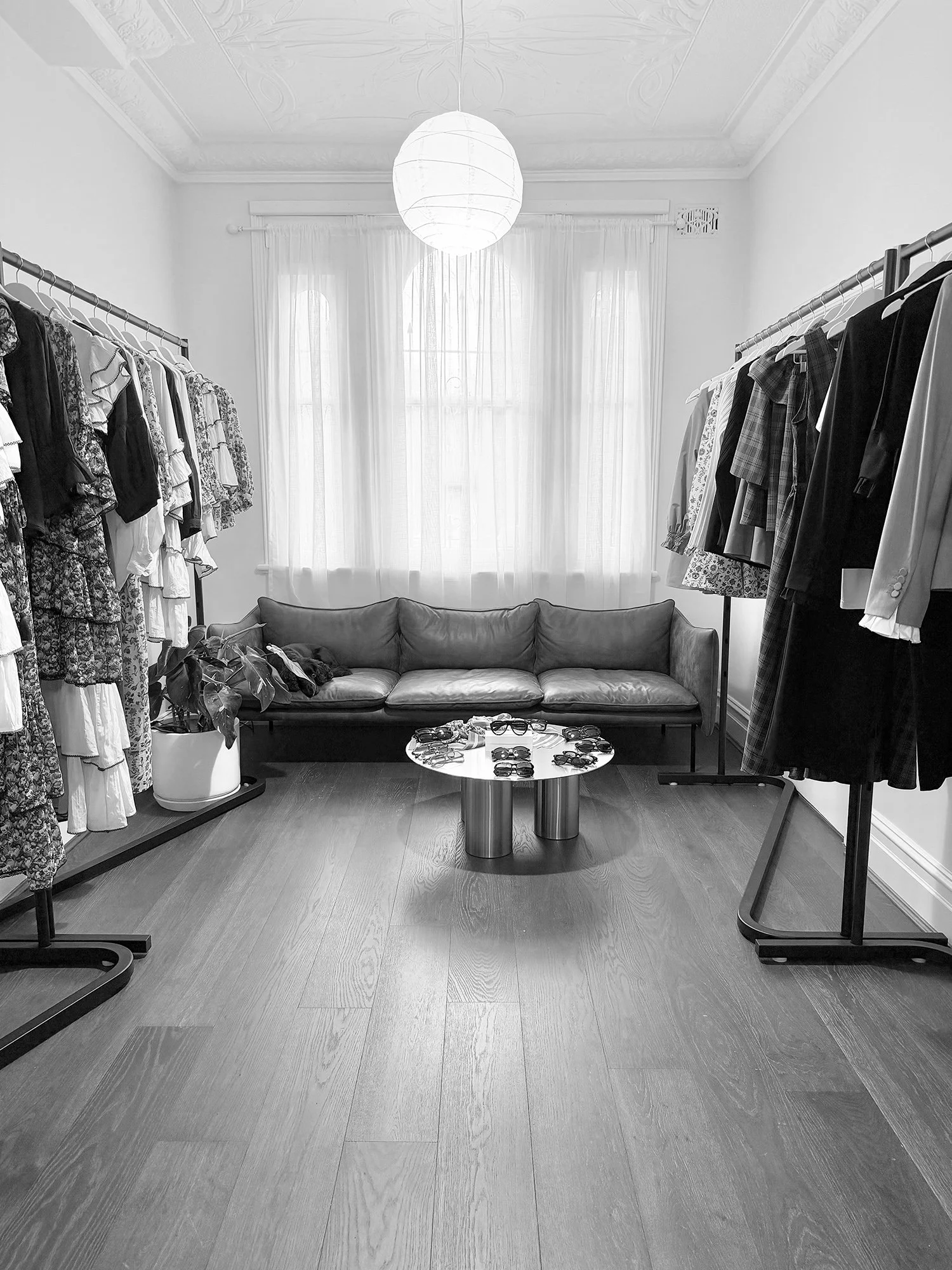 A room with white walls and ceiling, featuring ornate crown molding and a paper lantern ceiling light. There are clothing racks on both sides with various garments. A gray sofa is against the back wall under tall, sheer curtains covering large windows. A coffee table in front of the sofa holds numerous sunglasses, and a potted plant is near the left clothing rack.