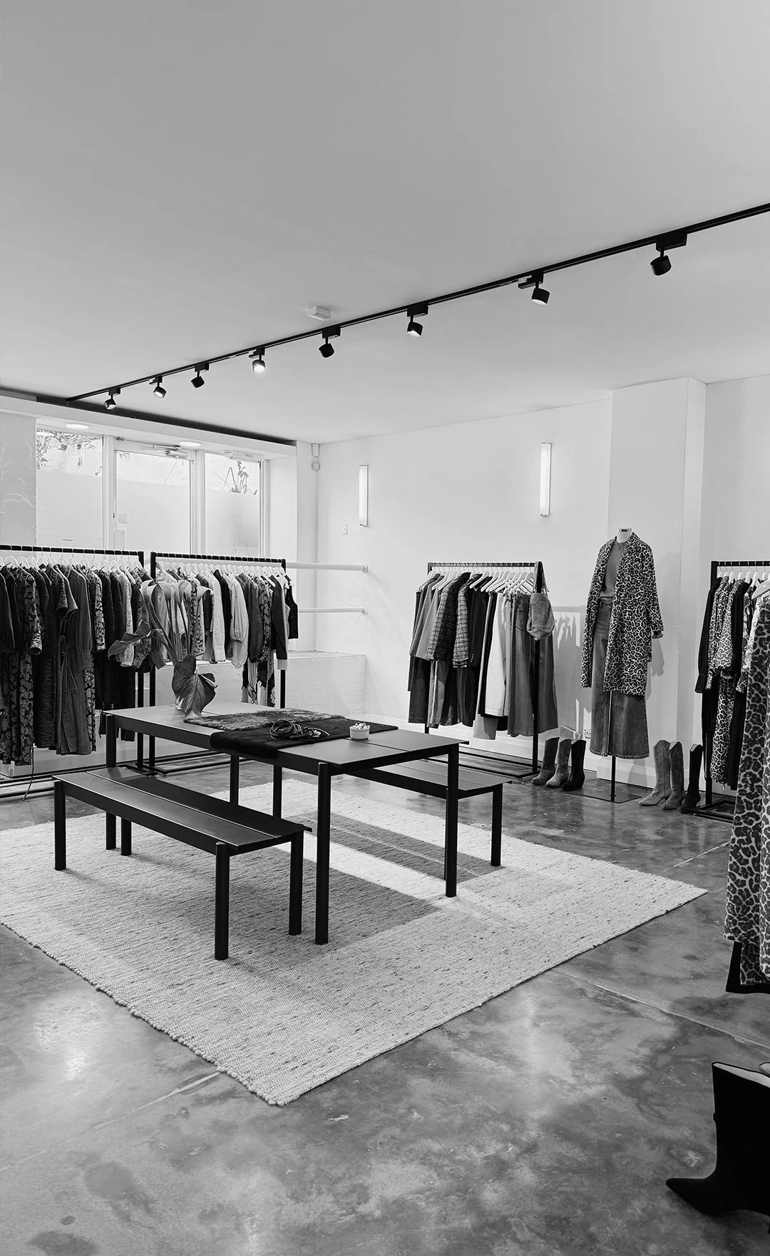 Black and white photo of a LTD Agency with racks of clothes, a mannequin wearing a leopard print coat, and a table with a plant and accessories, inside a modern store with a rug and track lighting.