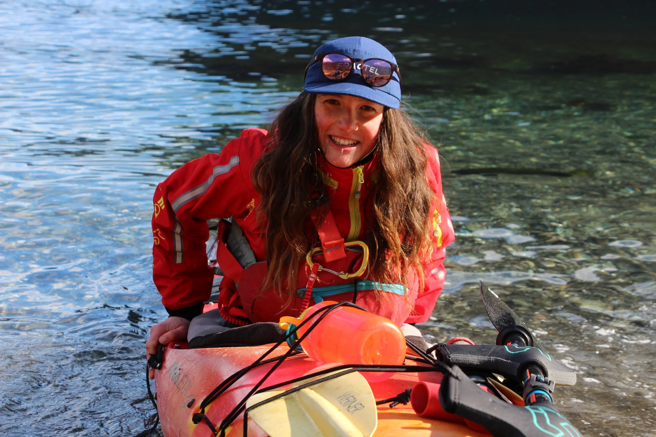 Woman in red jacket and blue hat smiling on kayak near shoreline with clear water and pebbles.