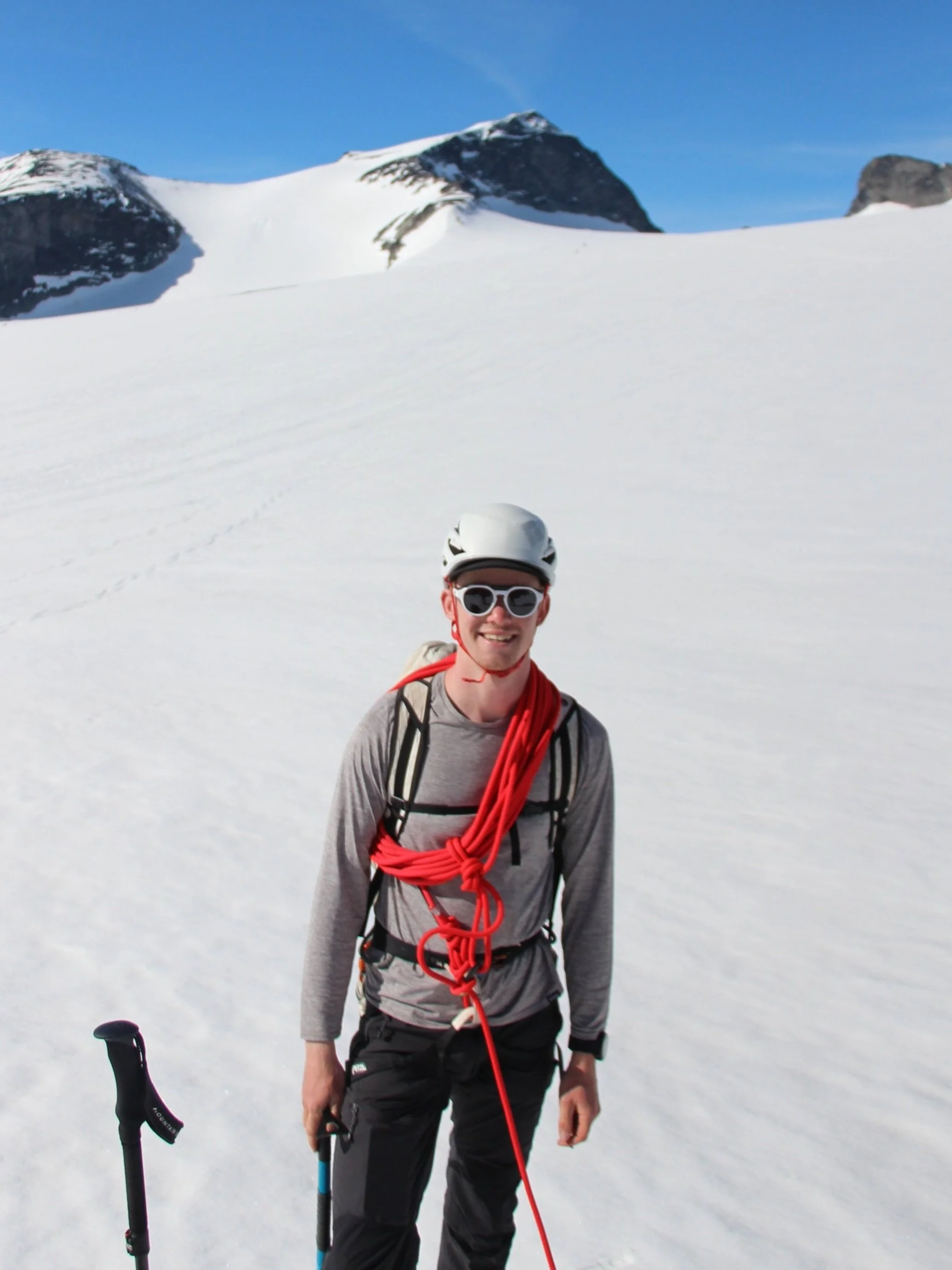 A person standing on snow in a mountainous area with snow-covered peaks in the background. The person is wearing a helmet, sunglasses, a backpack, and winter outdoor gear, holding an ice axe, with a red rope around their shoulders.