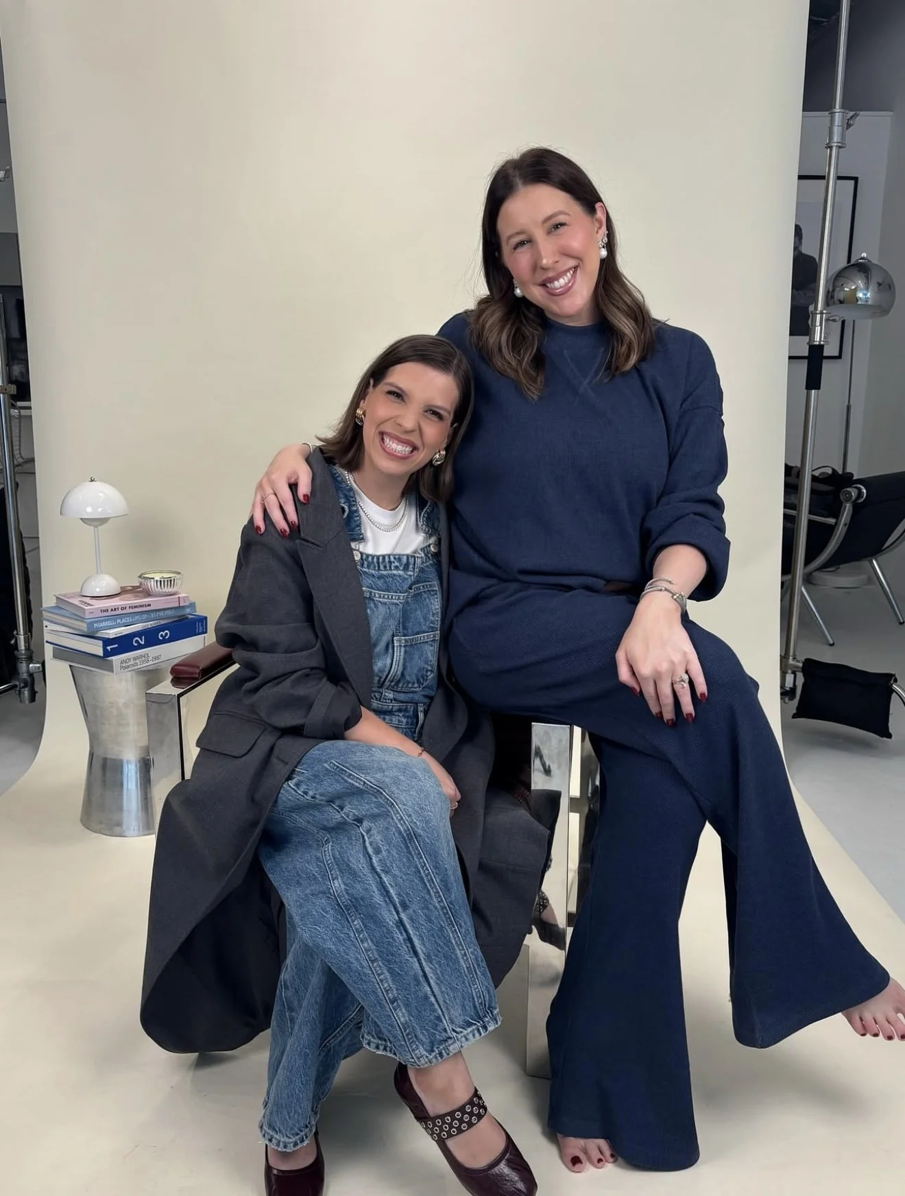 Two women sitting on a stool and a chair, smiling at the camera, in a studio setting with books, a lamp, and a light reflector in the background.