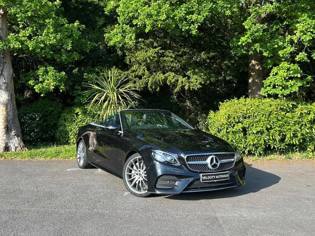 Black Mercedes-Benz convertible parked on an asphalt lot with trees and green bushes in the background