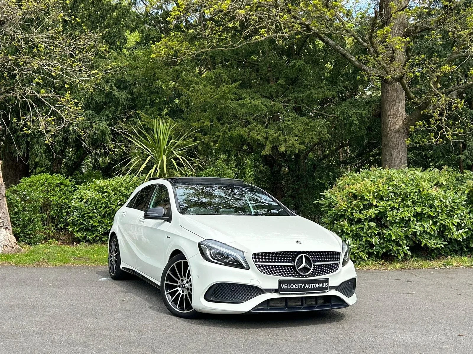 White Mercedes-Benz hatchback parked on a paved lot near green bushes and trees.