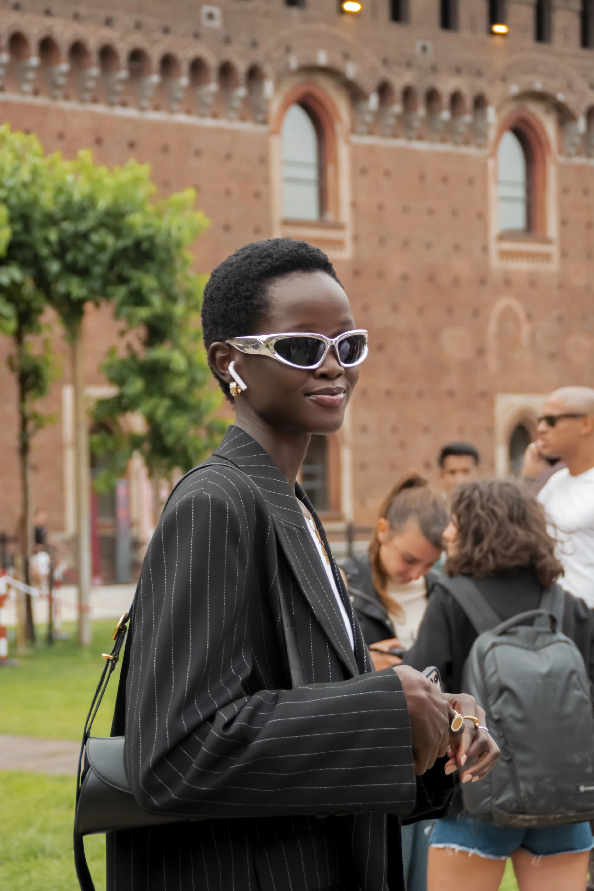 A stylish person wearing sunglasses, a pinstripe blazer, and carrying a shoulder bag is standing outdoors. They are holding a phone and wearing earbuds. A brick building, trees, and people are visible in the background.