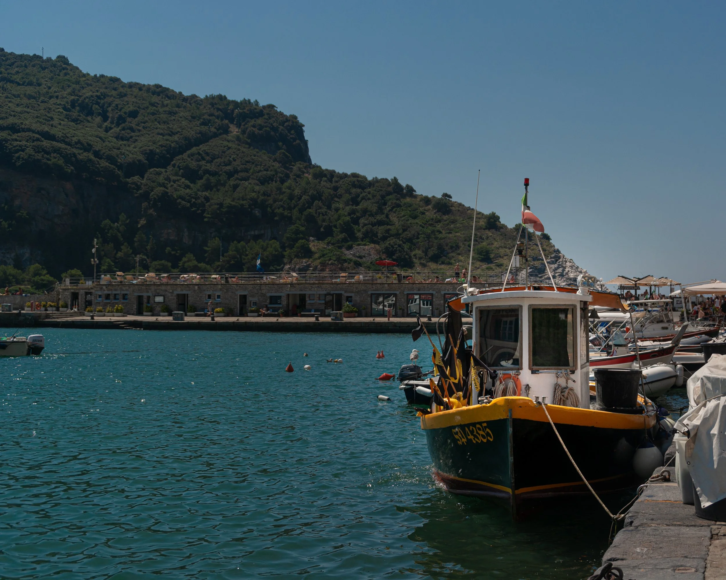 A small fishing boat docked at a marina with a mountainous landscape in the background, under a clear blue sky.