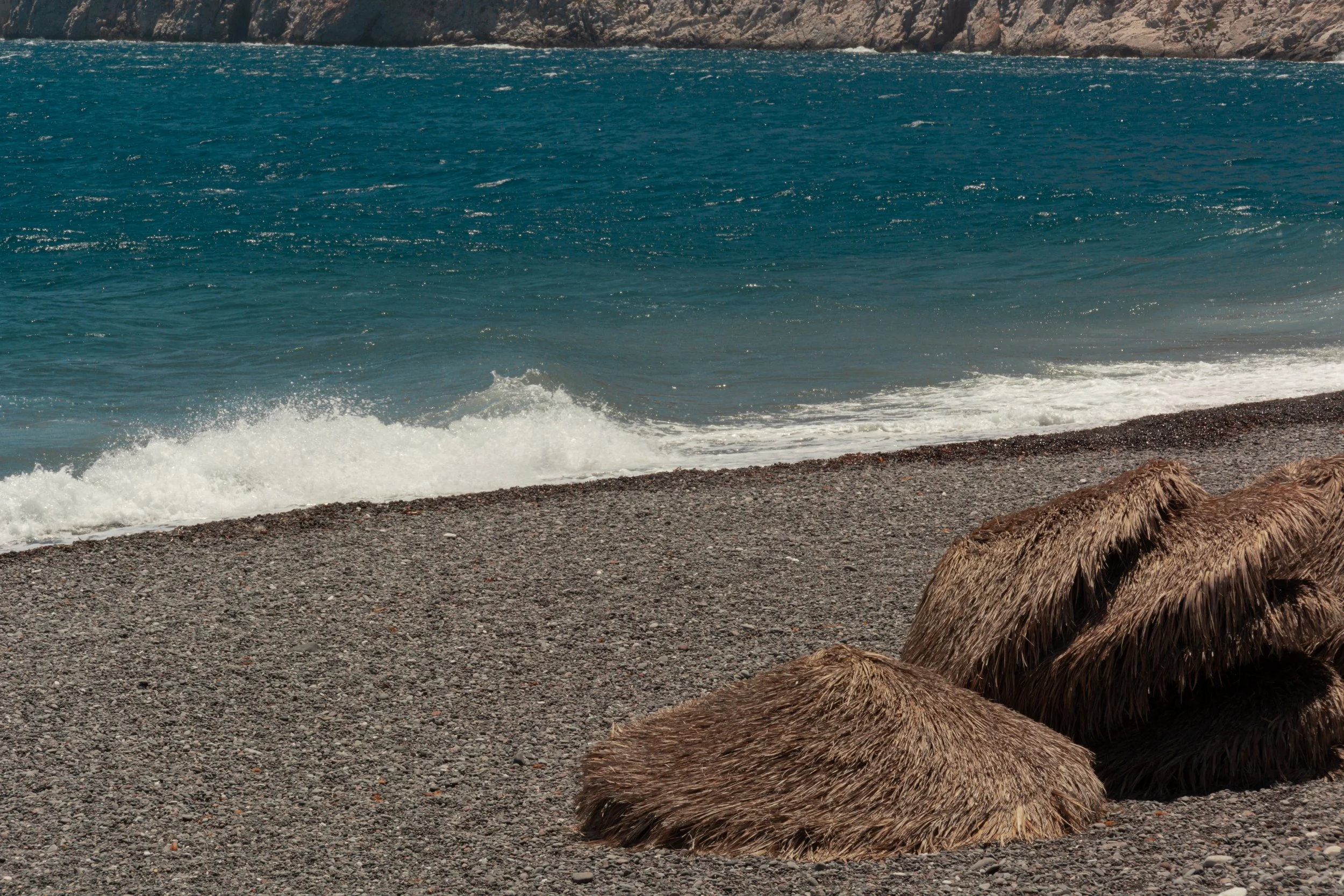Pebble beach with straw umbrellas, and ocean waves in the background.