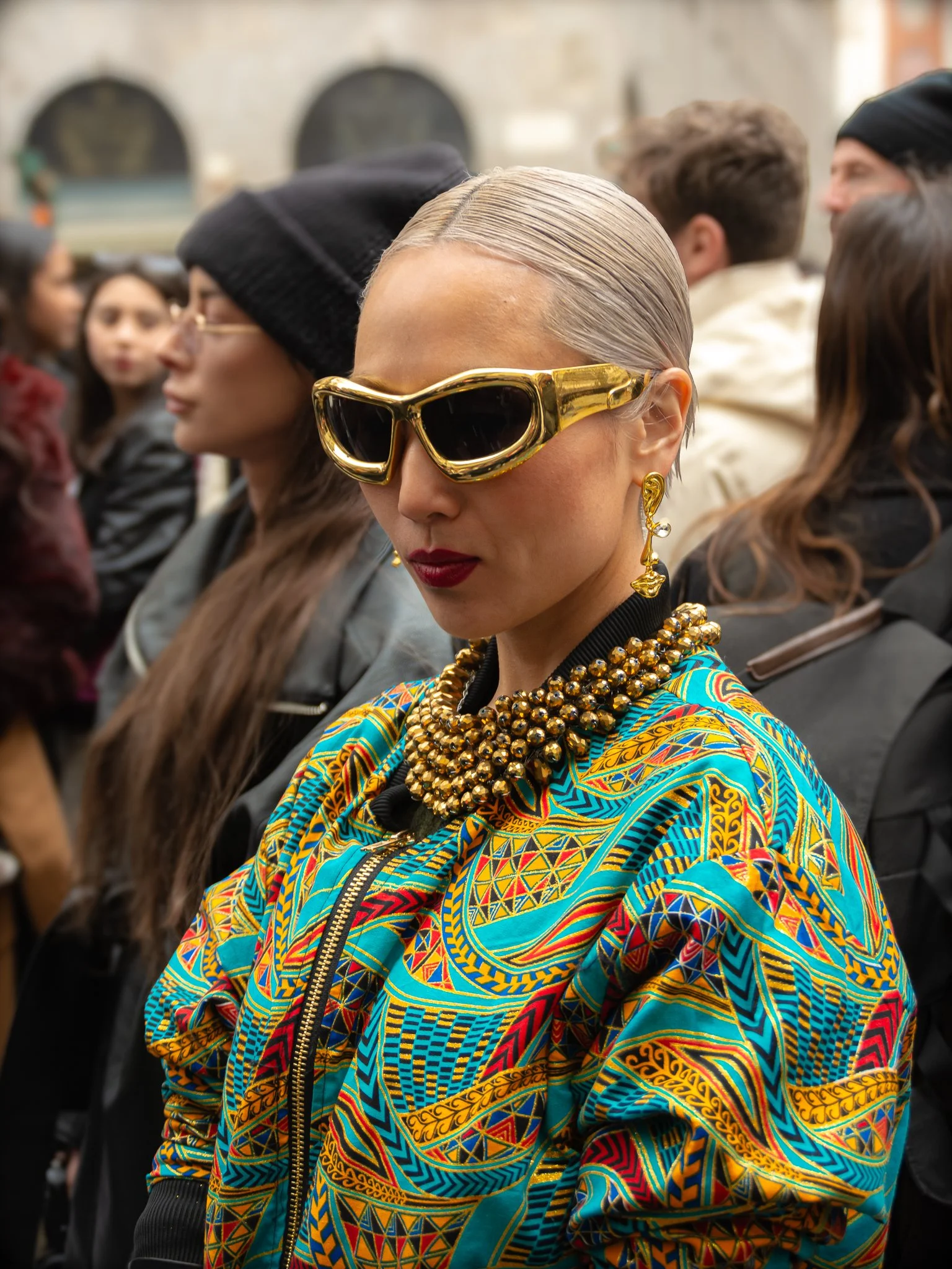 A fashionable Asian woman in a colorful geometric jacket, gold sunglasses, and accessories, is surrounded by the crowd on a street in Milan.