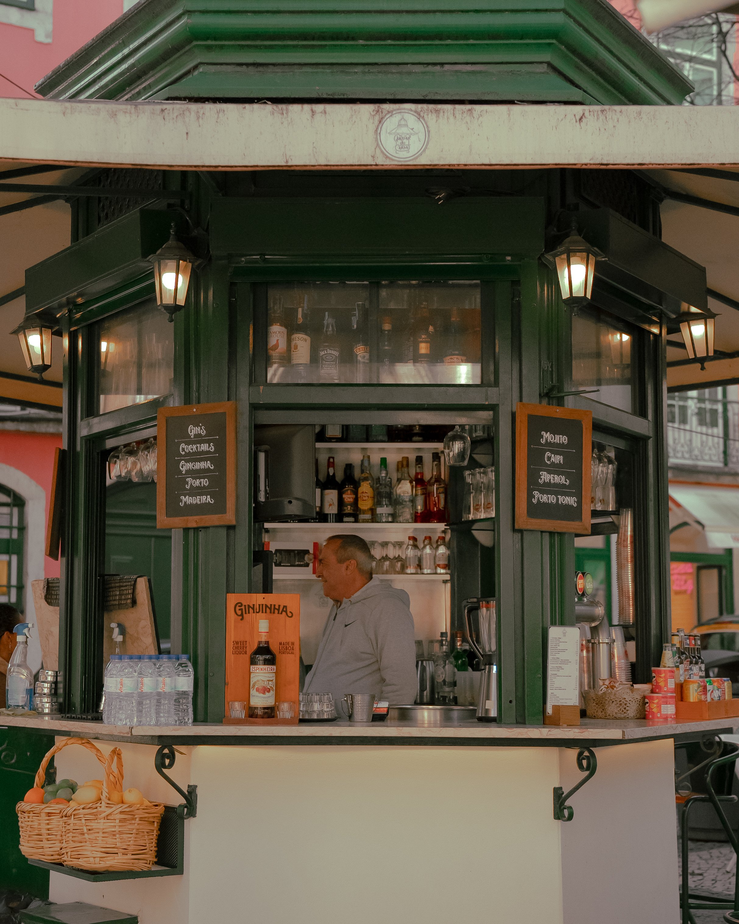 Outdoor kiosk bar with a bartender preparing drinks, featuring bottles and glasses on shelves, menu boards, beverage containers, and a fruit basket.