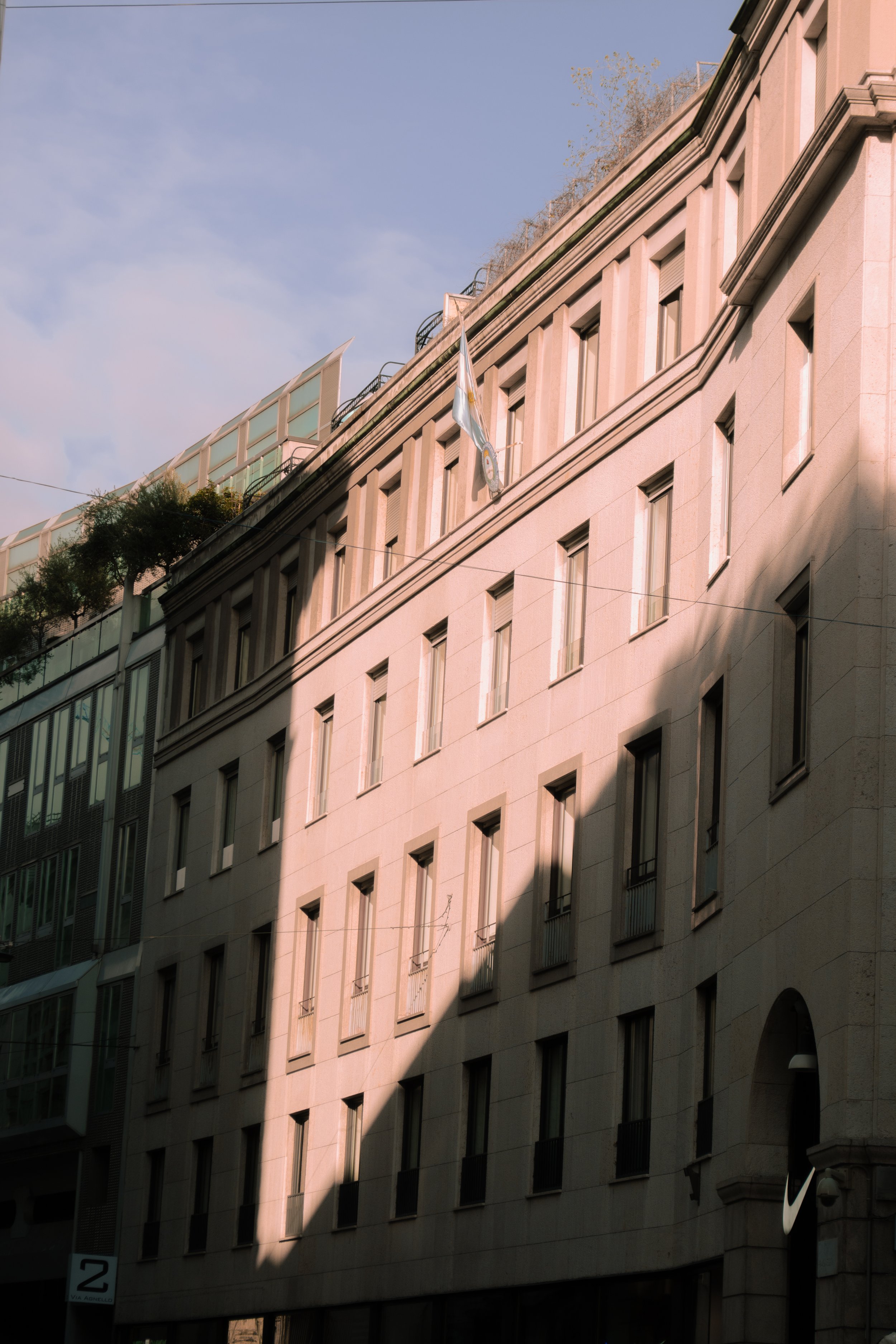 Facade of a beige stone building with numerous windows, a flag, and a partial shadow, adjacent to a modern glass structure.