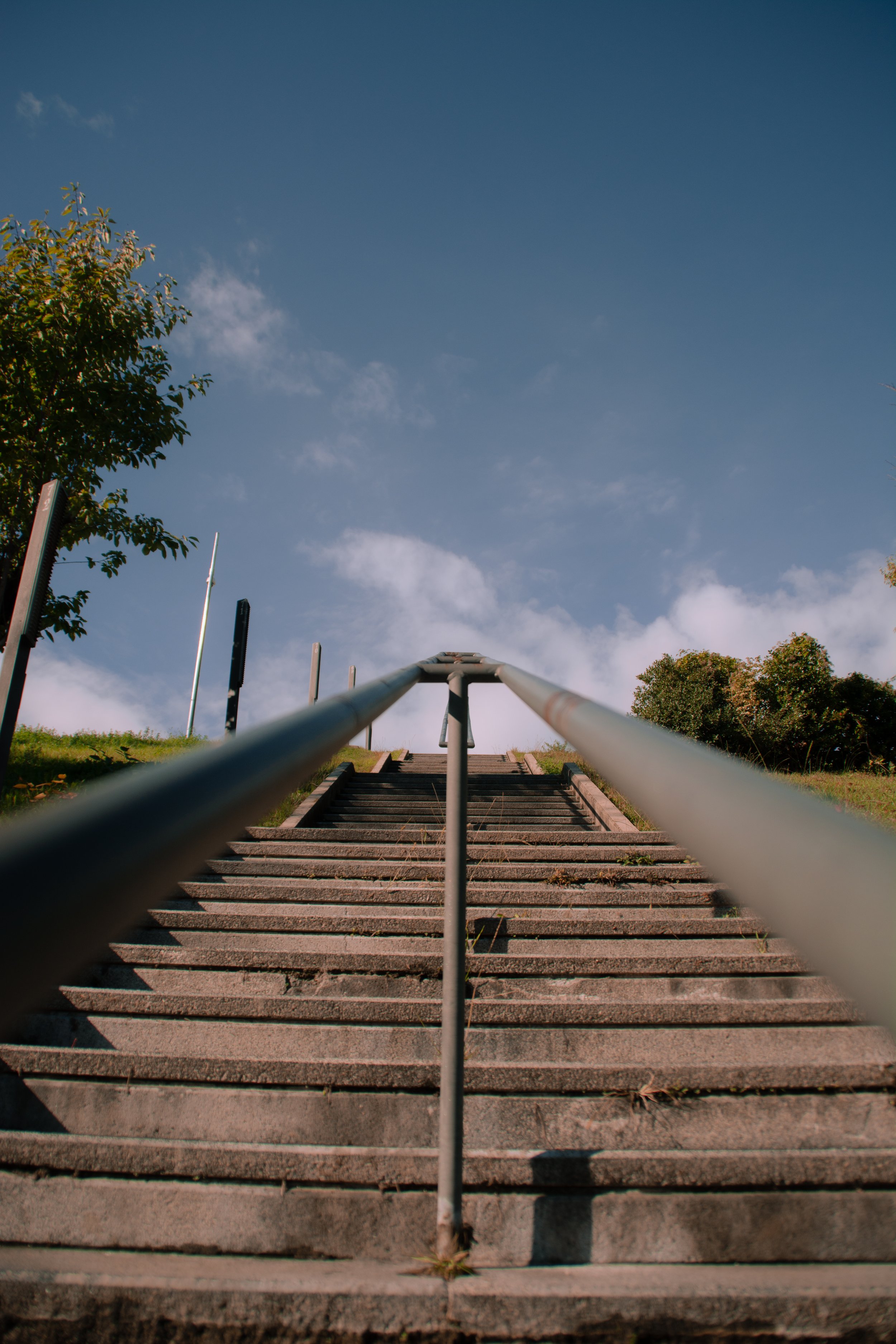 Concrete stairs with metal railing leading uphill, surrounded by greenery and under a blue sky with clouds.