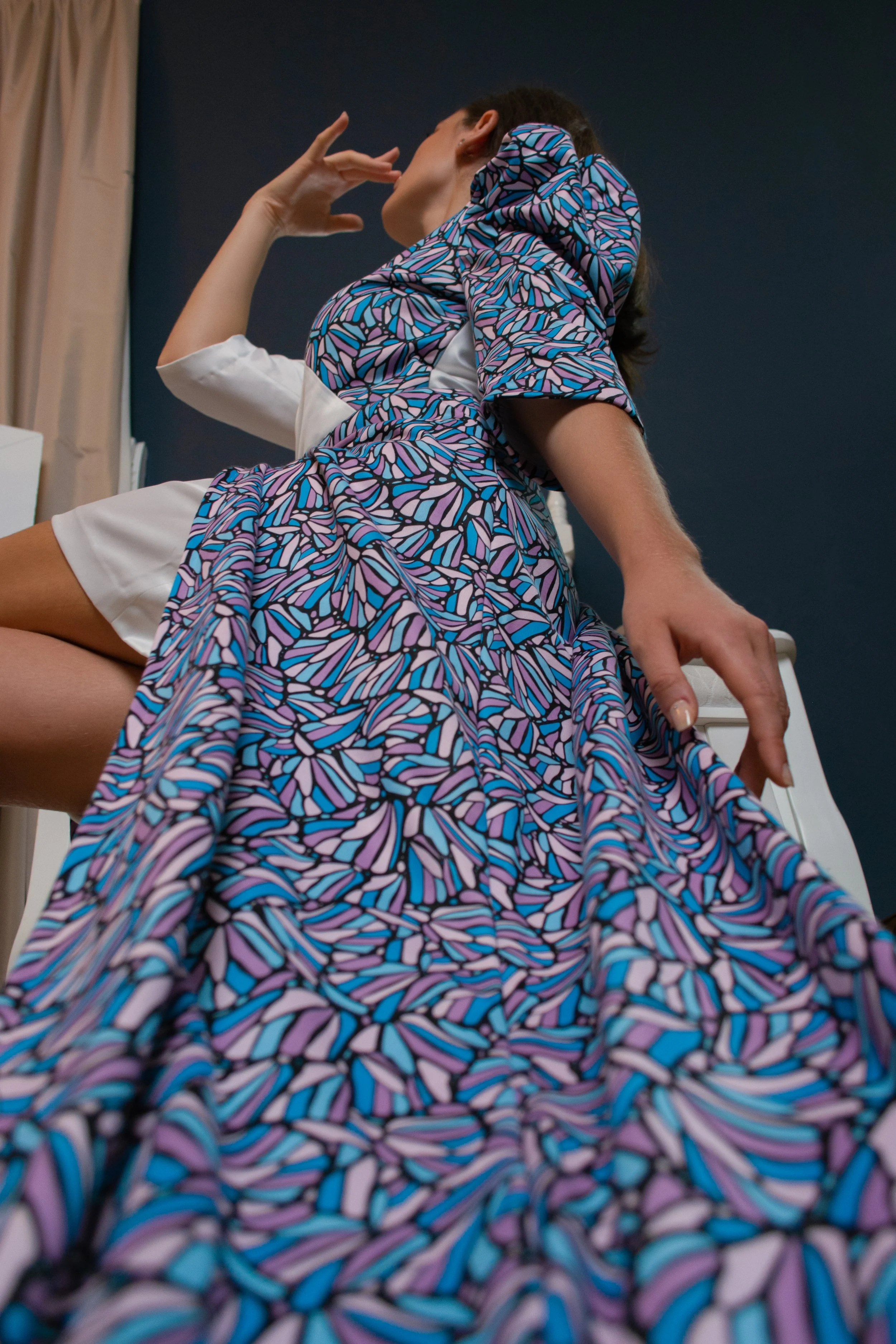 A model wearing a colorful dress with a geometric pattern, sitting in a stylish pose, with one hand extended by her side and the other one bent with the fingers grazing her face.