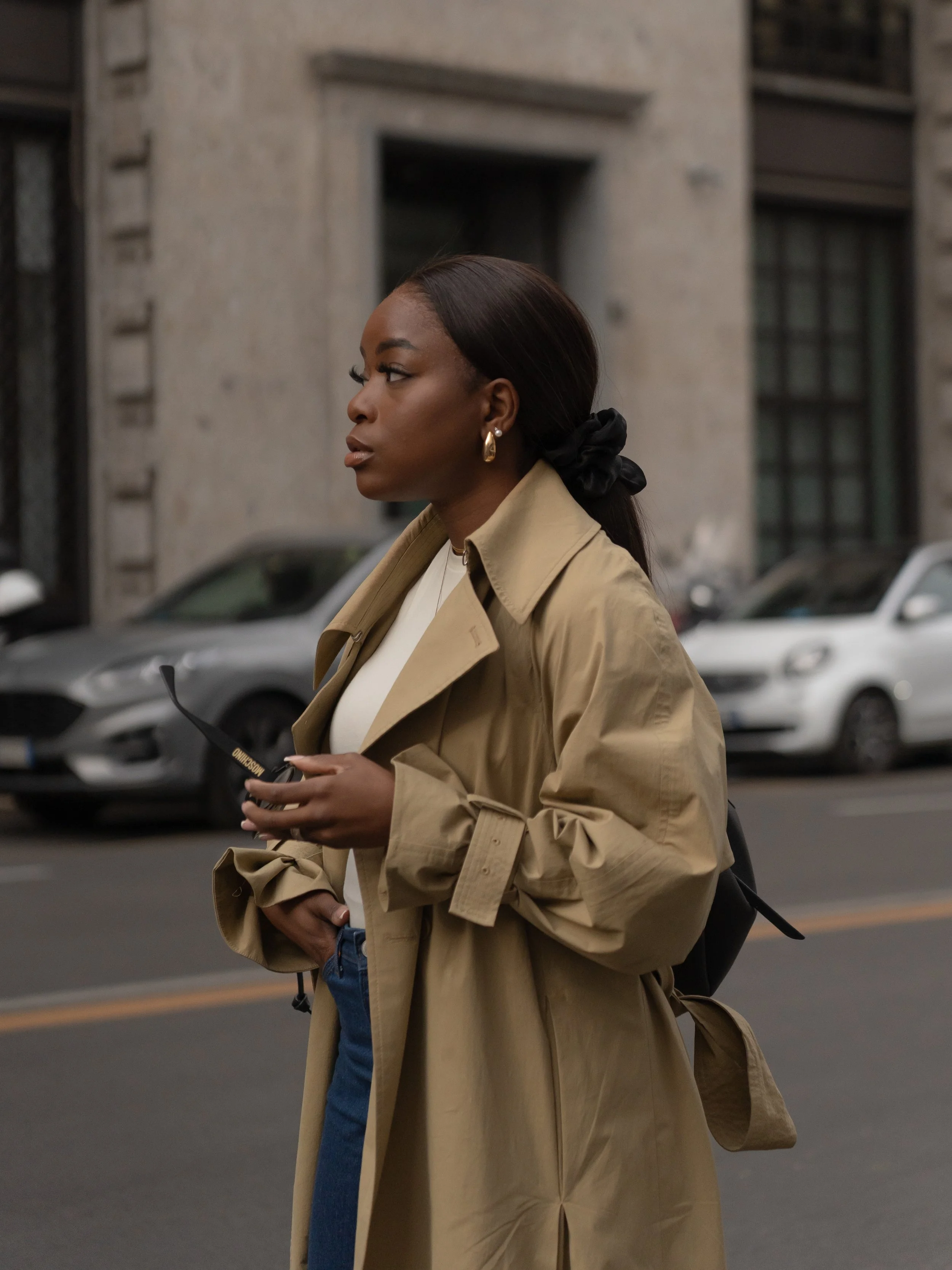 A woman in a beige trench coat, holding sunglasses, standing on the street.